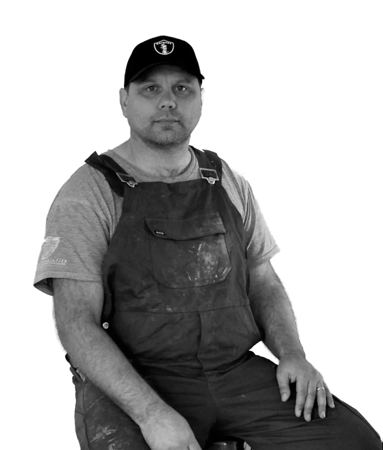A man wearing a black baseball cap and a dirty apron, sitting with his hands resting on his lap, in front of a plain background.