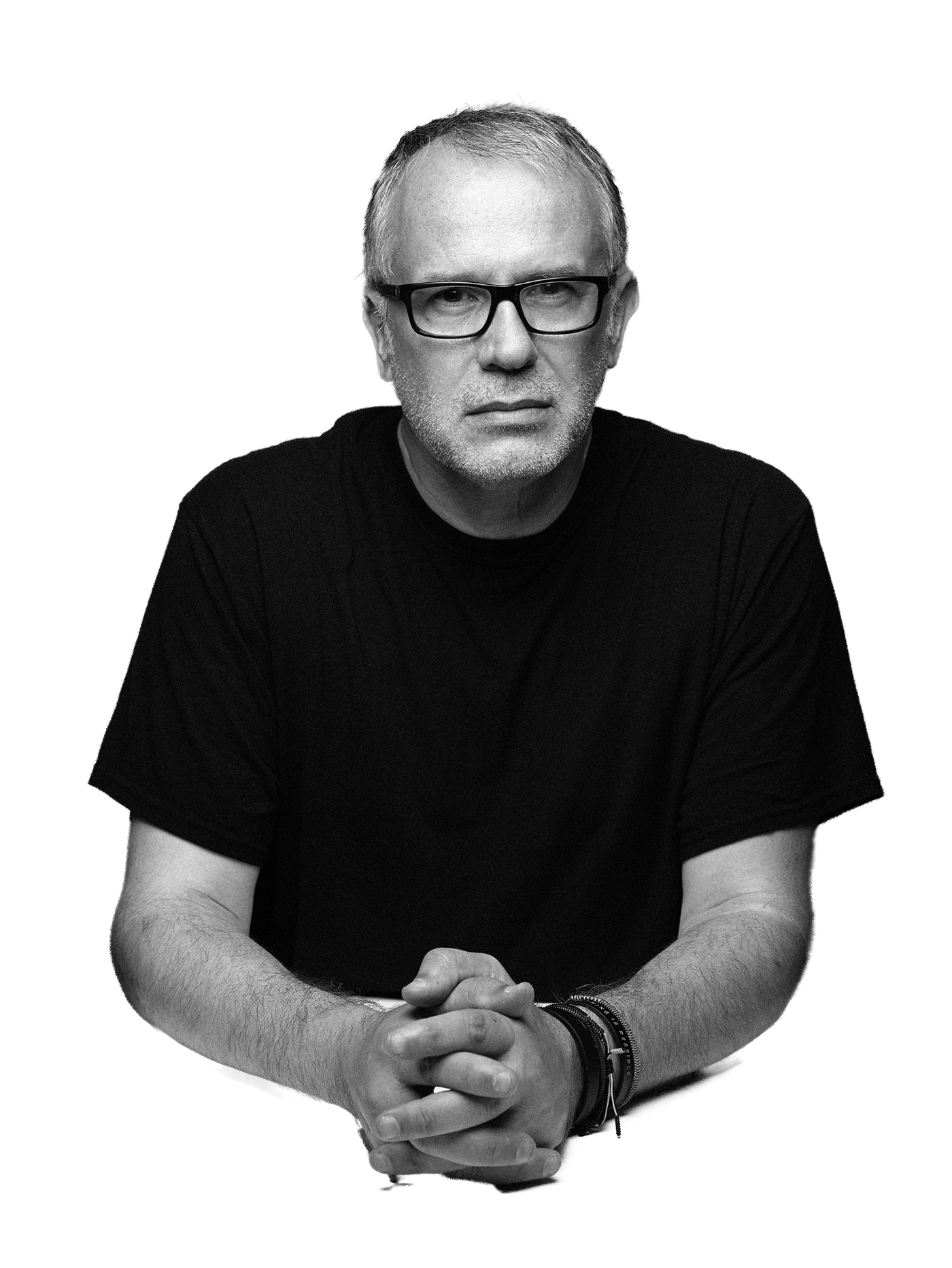 A black-and-white photo of a middle-aged man with glasses, short gray hair, and a beard, wearing a black t-shirt, sitting with clasped hands, against a dark background.