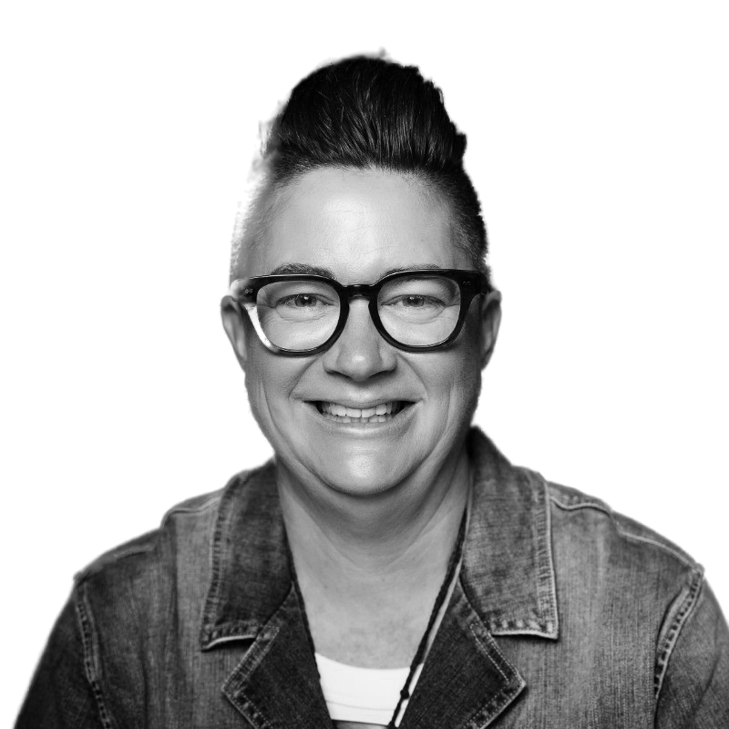 A smiling person with short dark hair styled upwards, wearing round black glasses and a denim jacket, against a black background.