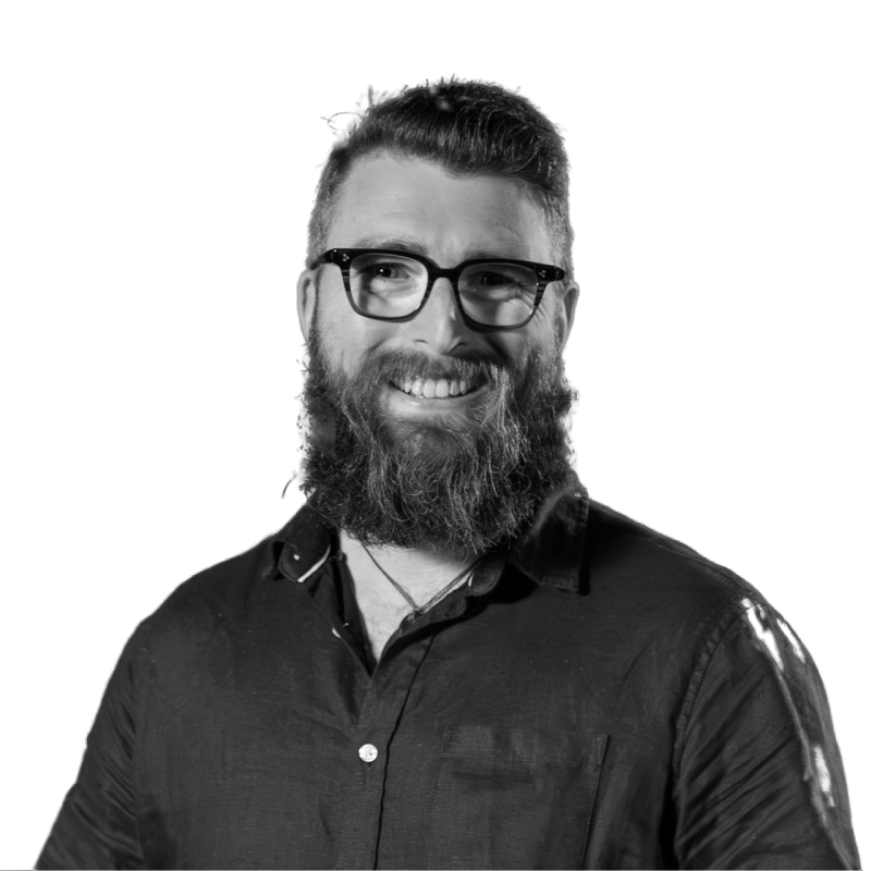 Black and white portrait of a smiling man with glasses and a beard, wearing a dark shirt, against a dark background.