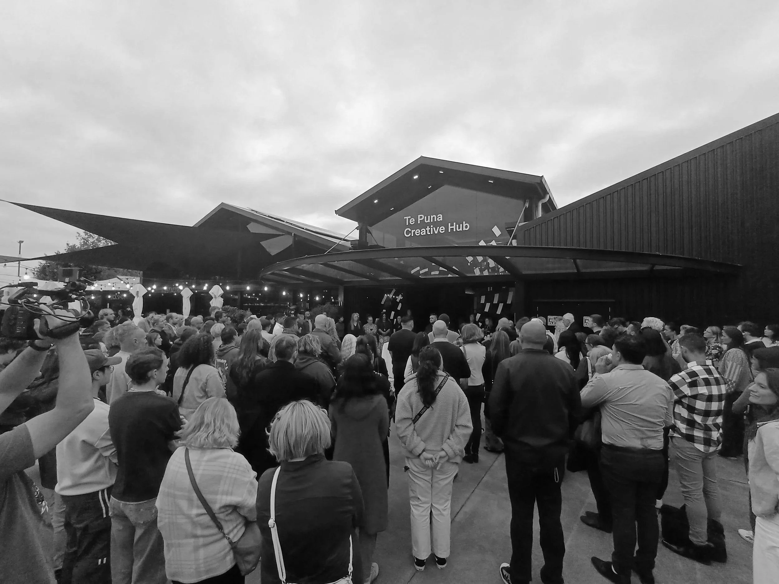 Crowd gathered outside the Te Puna Creative Hub building, with people standing and socializing beneath a cloudy sky.