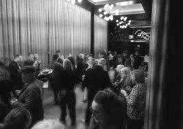 People mingling at a social event in a large room with curtains and a chandelier.