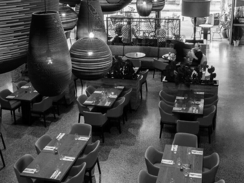 Empty restaurant with several tables set with glasses and napkins, large pendant lights hanging from ceiling, and a few patrons seated in the background.