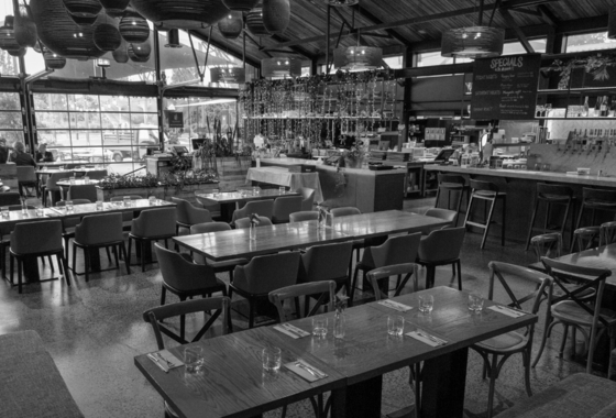 Empty restaurant or cafe with tables and chairs, large windows, and a bar area in the background.