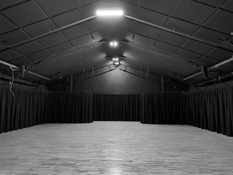 Empty indoor stage with wooden floor, black curtains, and overhead lighting in a large hall or theater.