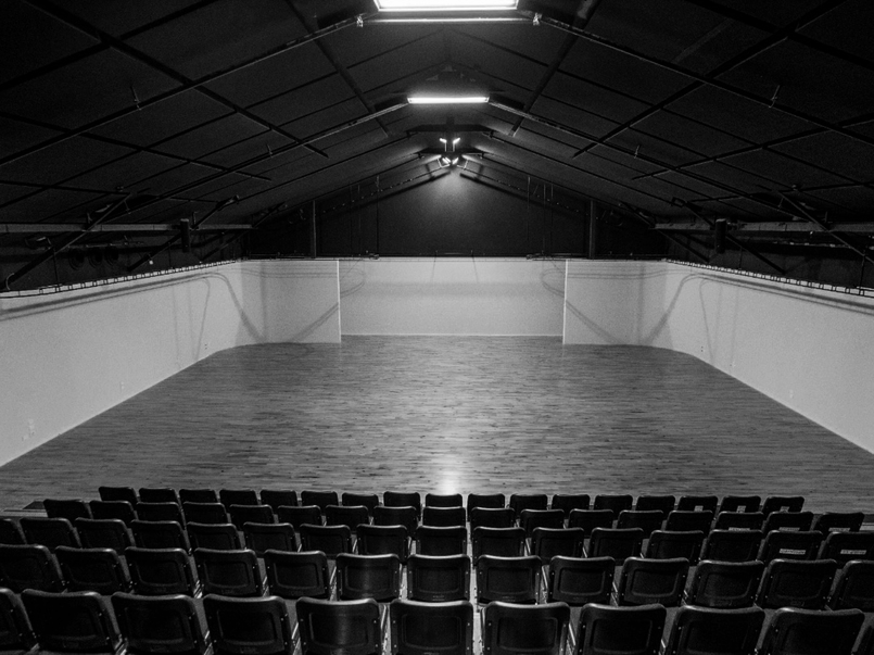 Empty indoor basketball court with wooden flooring, white walls, and black folding chairs in the foreground.