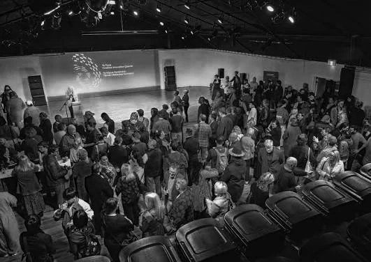 Crowd of people gathered in a large indoor event space with a stage and projection screen