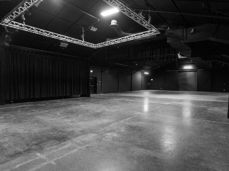 Empty black box theater with a wide stage, black curtains on the side, and stage lighting rig above.
