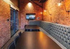 Interior view of a modern dining area with a brick wall, a long tufted gray banquette, black dining table, and wall-mounted lights.