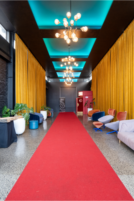 Long hallway with a red carpet, golden curtains on both sides, modern chairs, potted plants, and hanging chandeliers with exposed bulbs, ceiling lighting with teal panels.