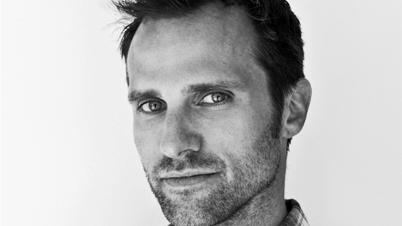 Close-up black and white portrait of a man with short dark hair and light-colored eyes, looking directly at the camera with a neutral expression.