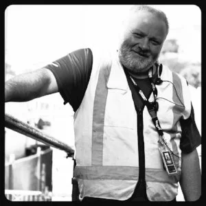 A man with a beard and short hair wearing a safety vest and a badge, smiling outdoors.