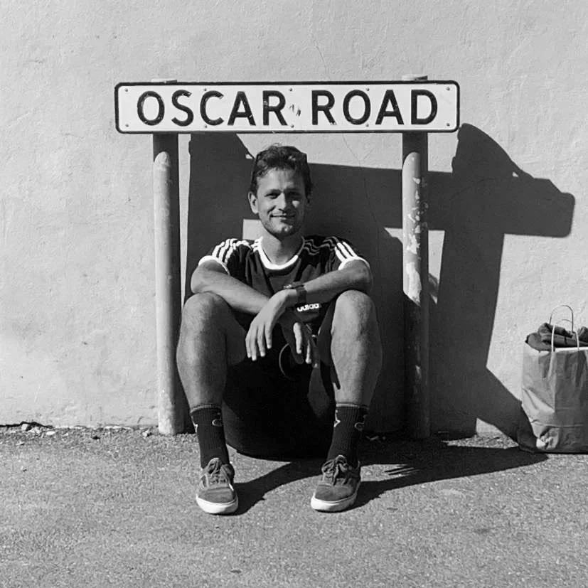 A young man sitting cross-legged on the ground in front of a street sign that says "Oscar Road." He is smiling and wearing a sports jersey, shorts, socks, and sneakers. There is a shadow of the sign on the wall behind him, and a bag is placed to his right.