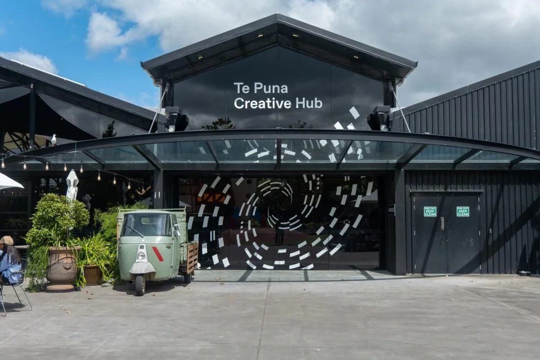Black building with a glass awning, white and black swirling mural, and sign reading 'Te Puna Creative Hub'.