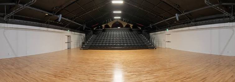 Empty indoor gymnasium or performance space with a wooden floor and tiered seating at one end.