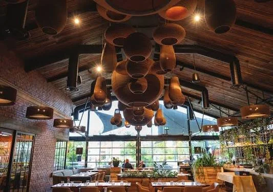 Interior of a restaurant or cafe with a wooden ceiling and unique hanging pendant lights, large windows, and seating arrangements.