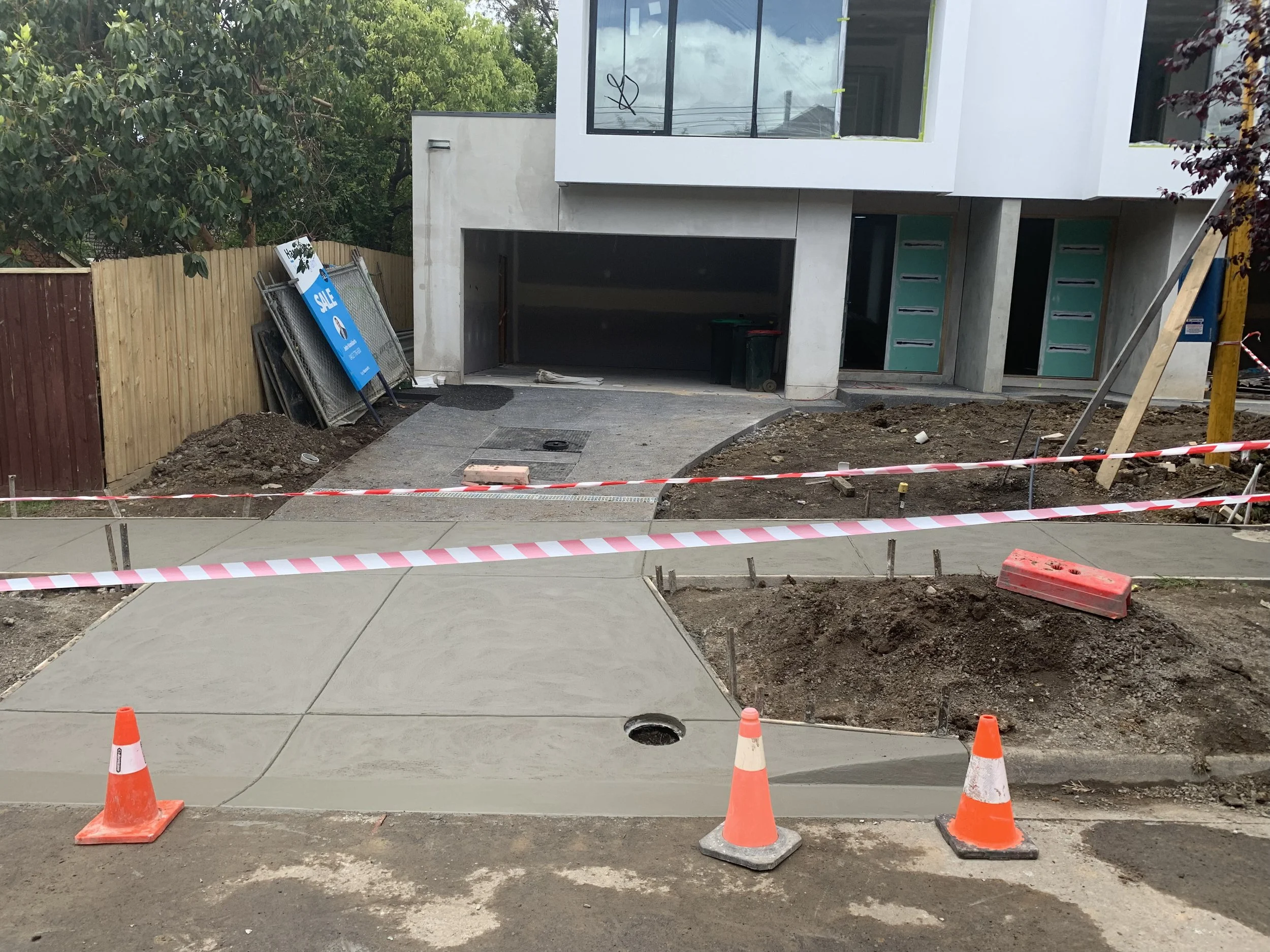 Construction site of a new residential building with a sidewalk being poured and marked with orange cones and caution tape, with an unfinished garage and partially completed exterior.