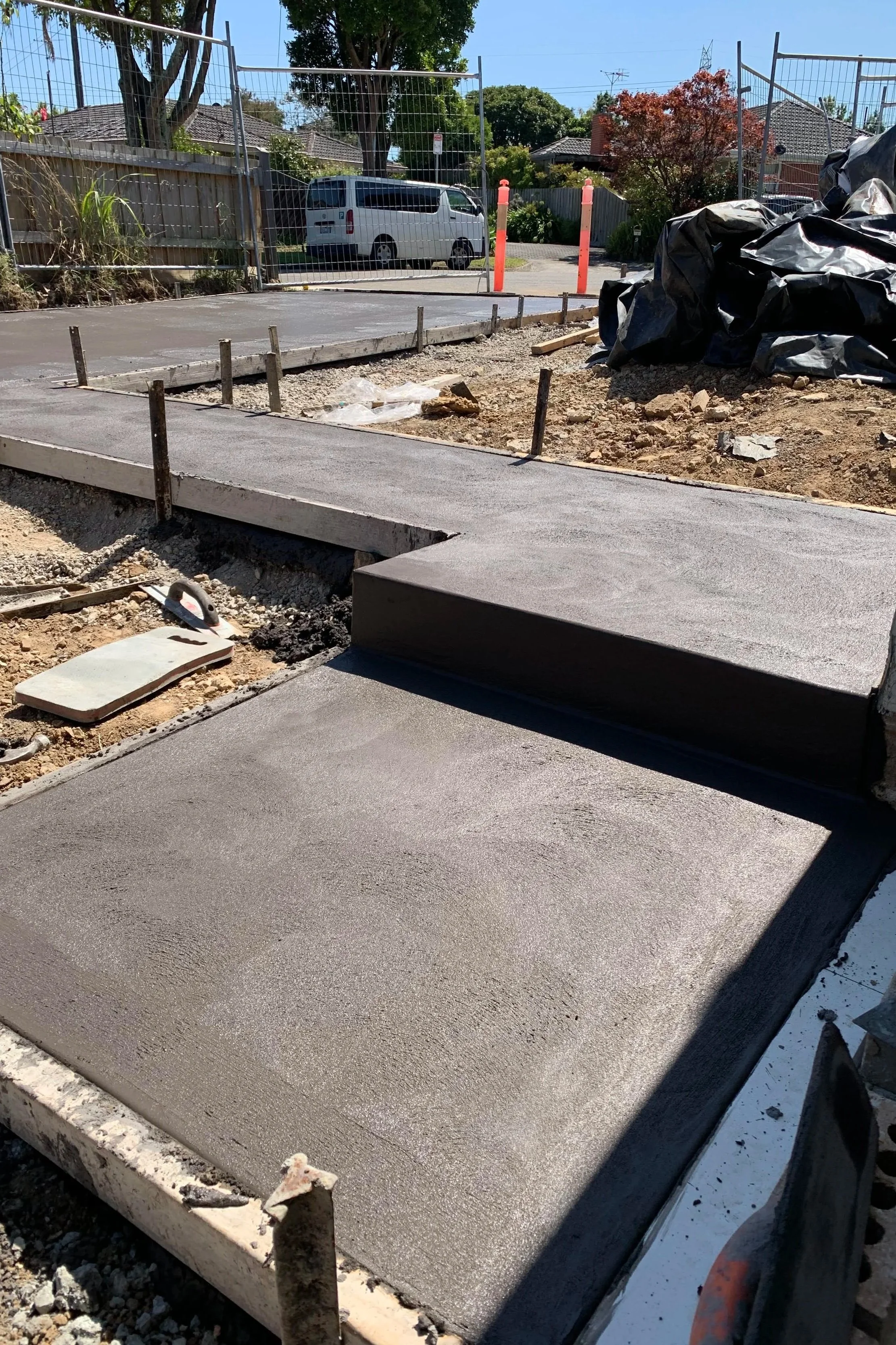 Concrete sidewalk construction with freshly poured concrete slabs, surrounded by construction tools, gravel, and barrier rods, in a residential neighborhood.