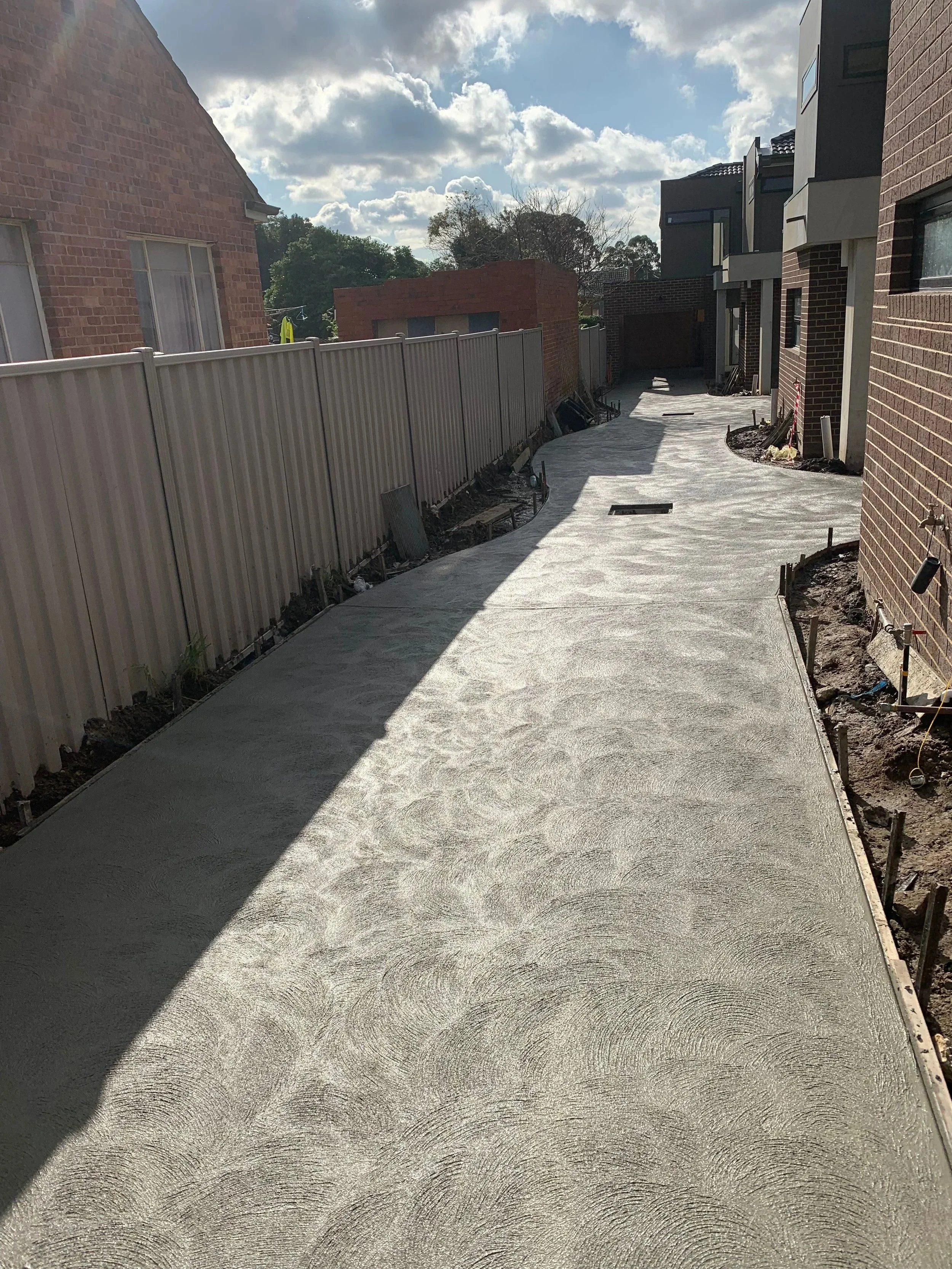 Recently paved concrete driveway in a residential backyard, surrounded by a beige metal fence on the left and brick walls of houses on the right, with construction tools and materials along the edges, and a partly cloudy sky overhead.