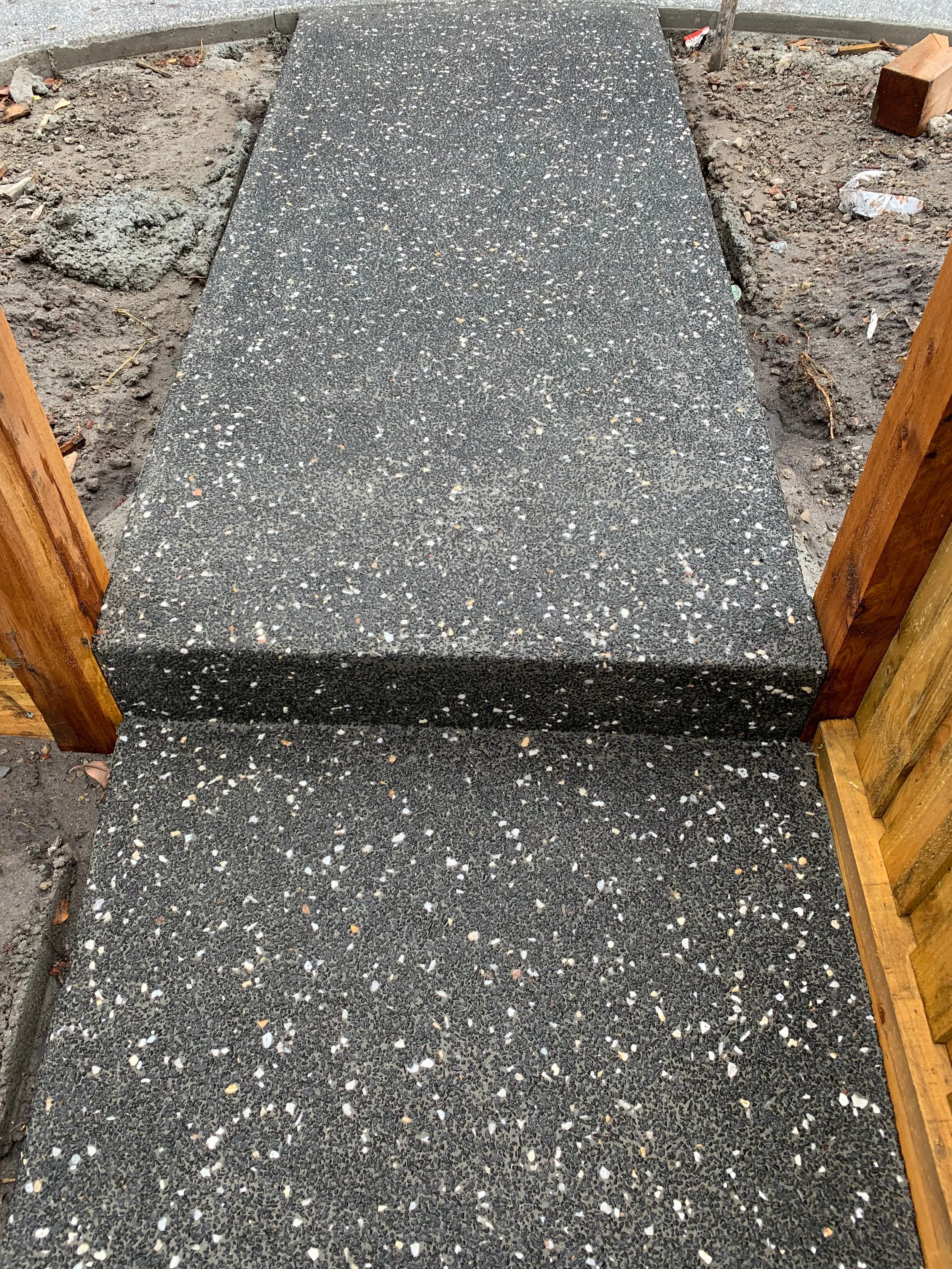 Close-up of a black speckled concrete step, part of a staircase under construction, with wooden framing on each side and dirt surrounding the area.