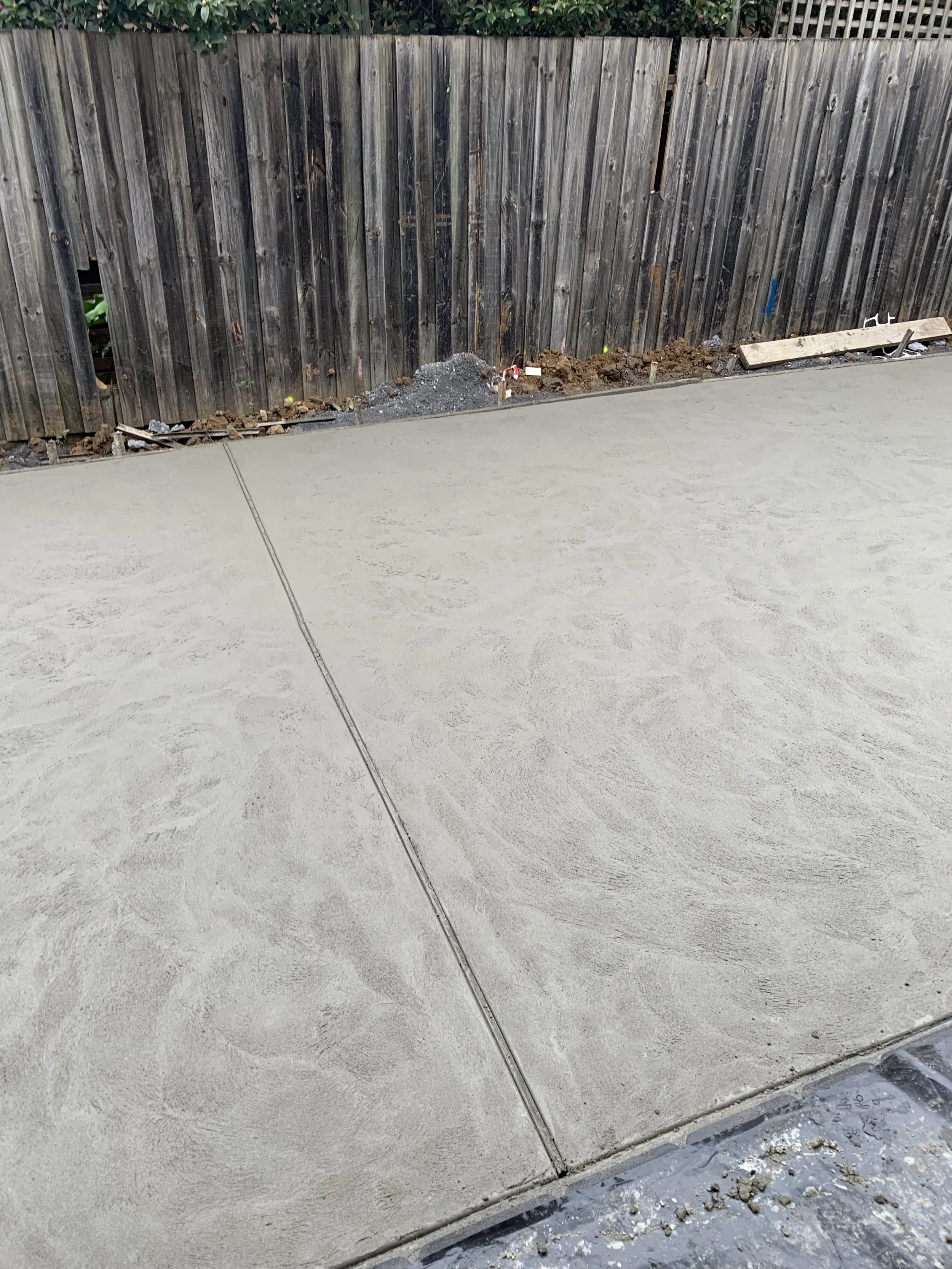 Newly poured concrete sidewalk with expansion joint, bordered by a wooden fence with some ongoing landscaping work.