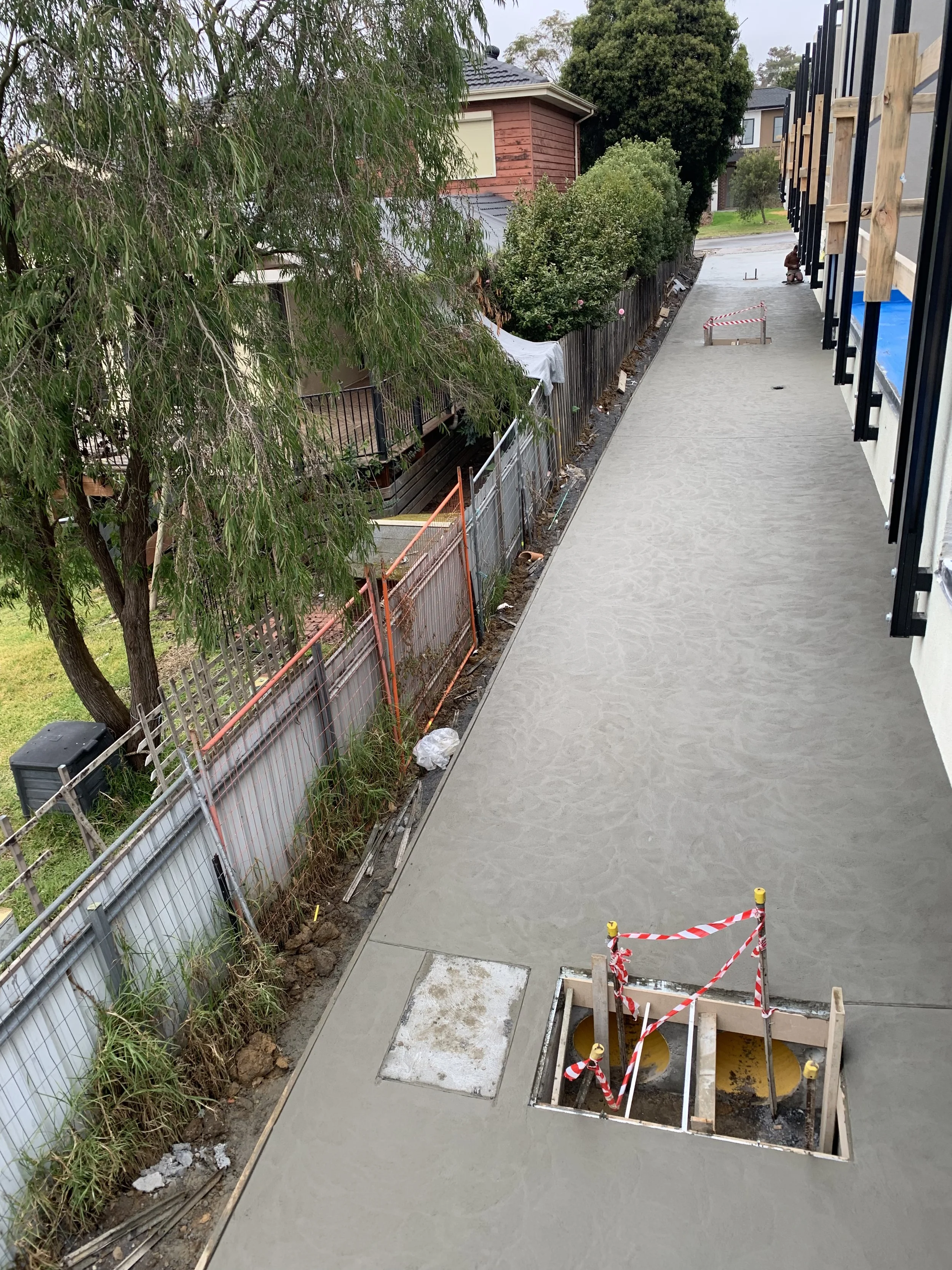 Unfinished sidewalk with open utility access points, construction barriers, and a fence along the building and residential area.