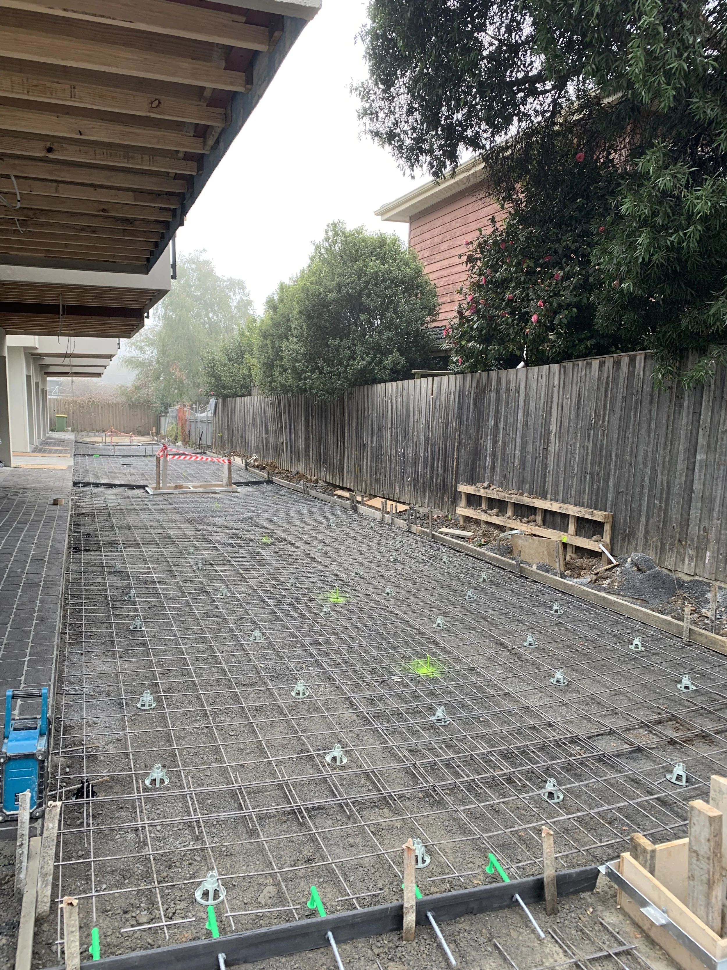 Construction site with a concrete foundation prepared with metal rebar for a new structure, with some green markings and construction barriers in the background, beside a wooden fence and trees.