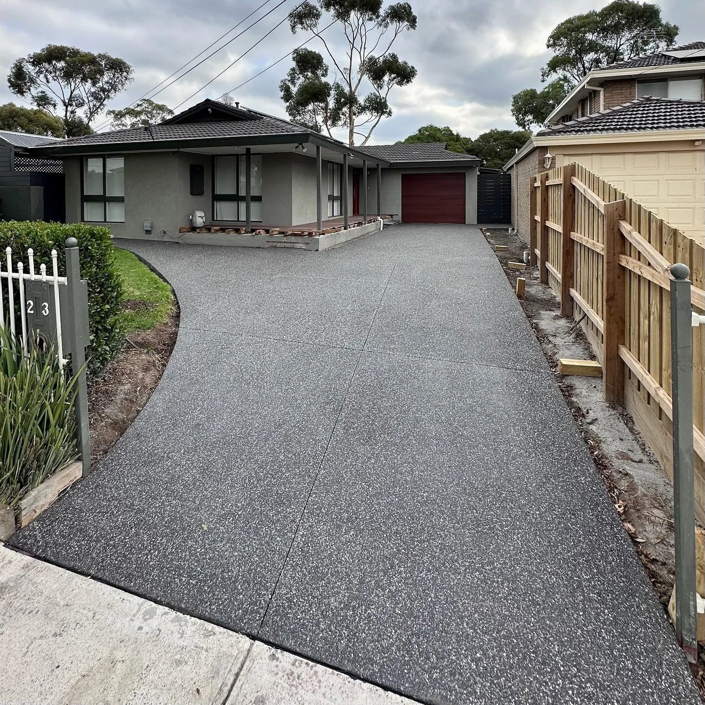 Newly paved driveway leading to a modern house with a garage, surrounded by a wooden fence on one side and a white fence on the other, with cloudy sky overhead.