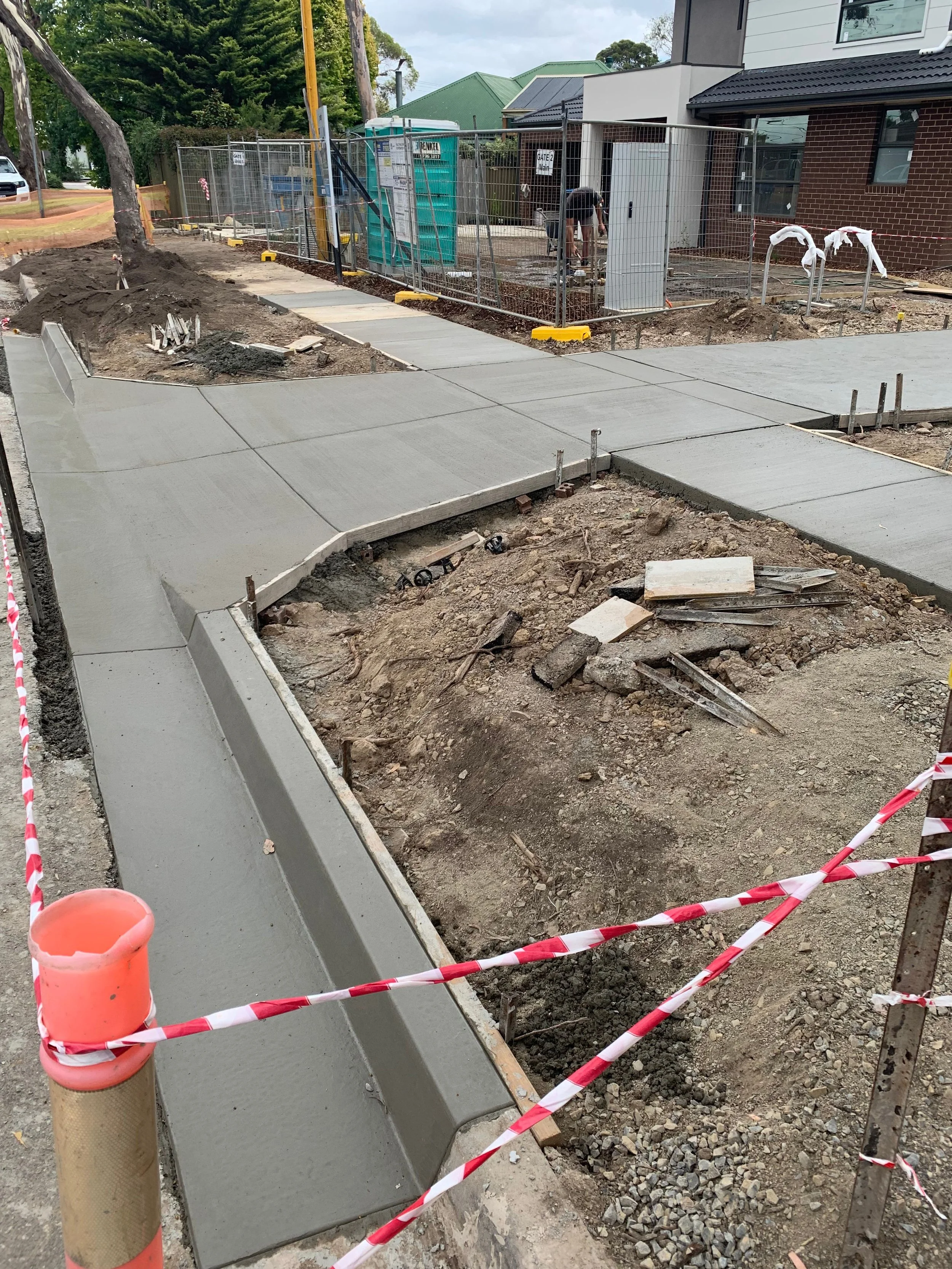 Construction site with newly poured concrete sidewalk and ongoing pavement work, fenced off with construction barriers and tools scattered around.