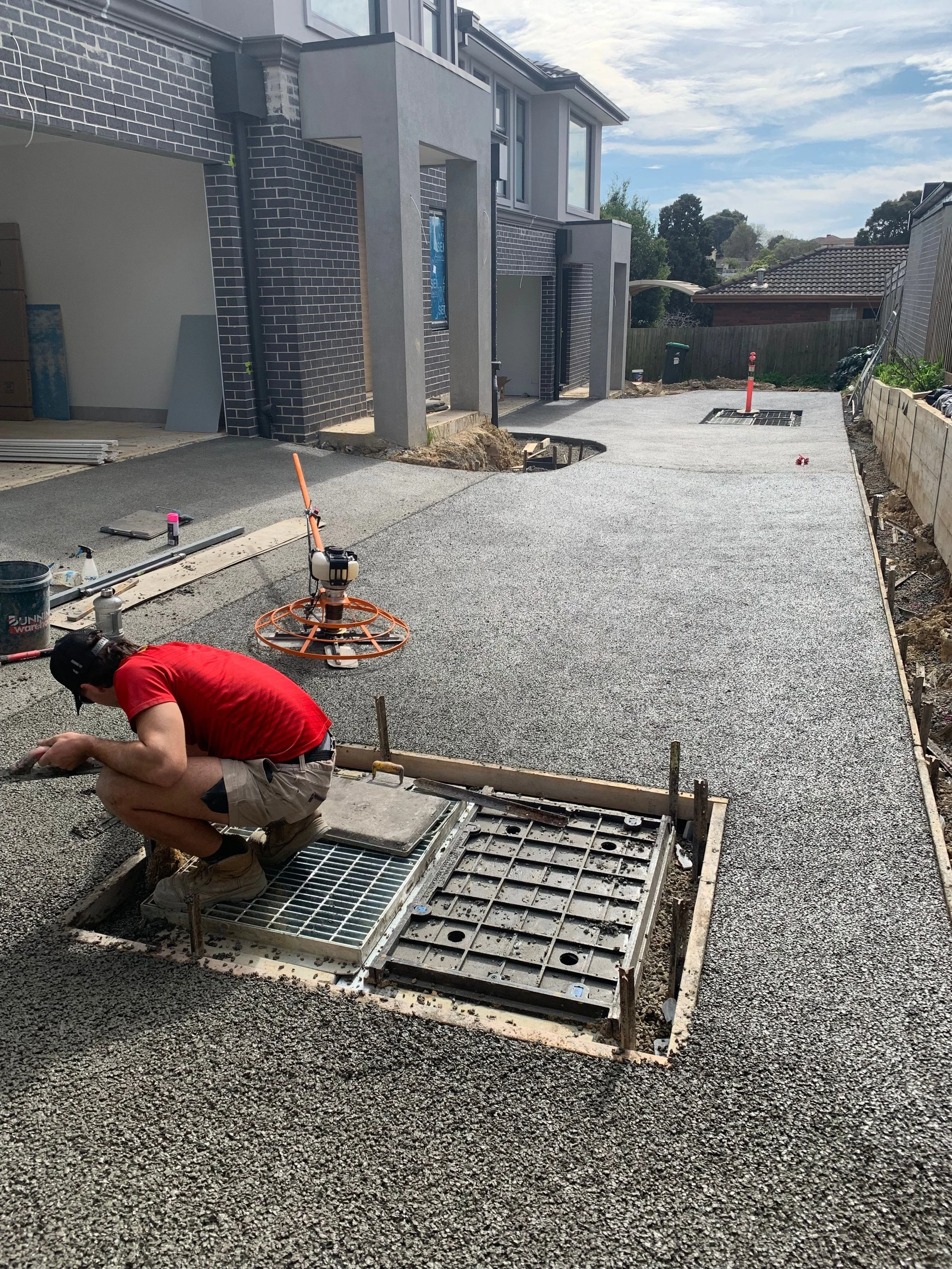 A construction worker crouching down and working on a manhole cover on a newly paved driveway in front of a modern house under construction.