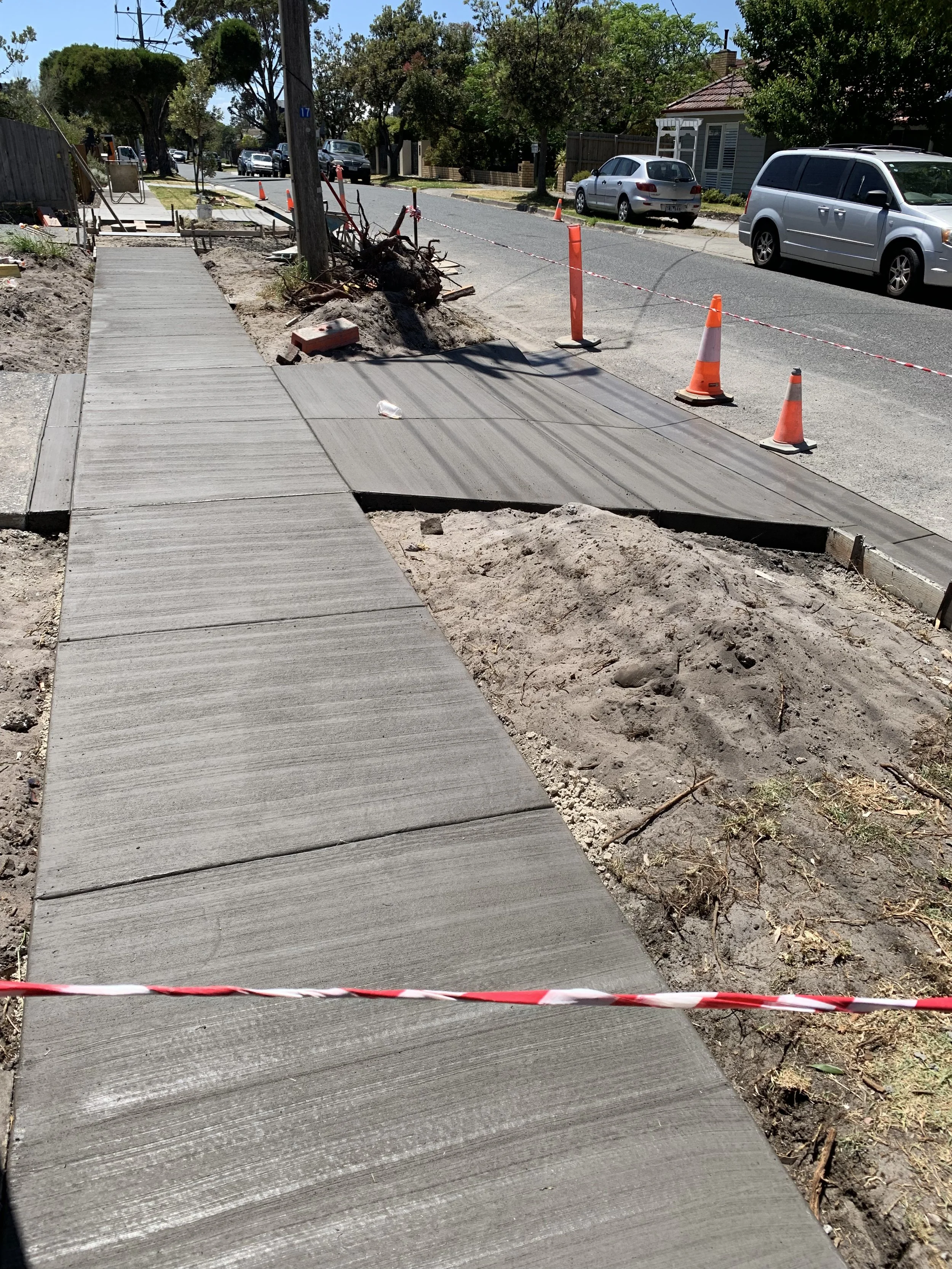 A sidewalk under construction with orange traffic cones and caution tape, partially paved, with some dirt and debris around.