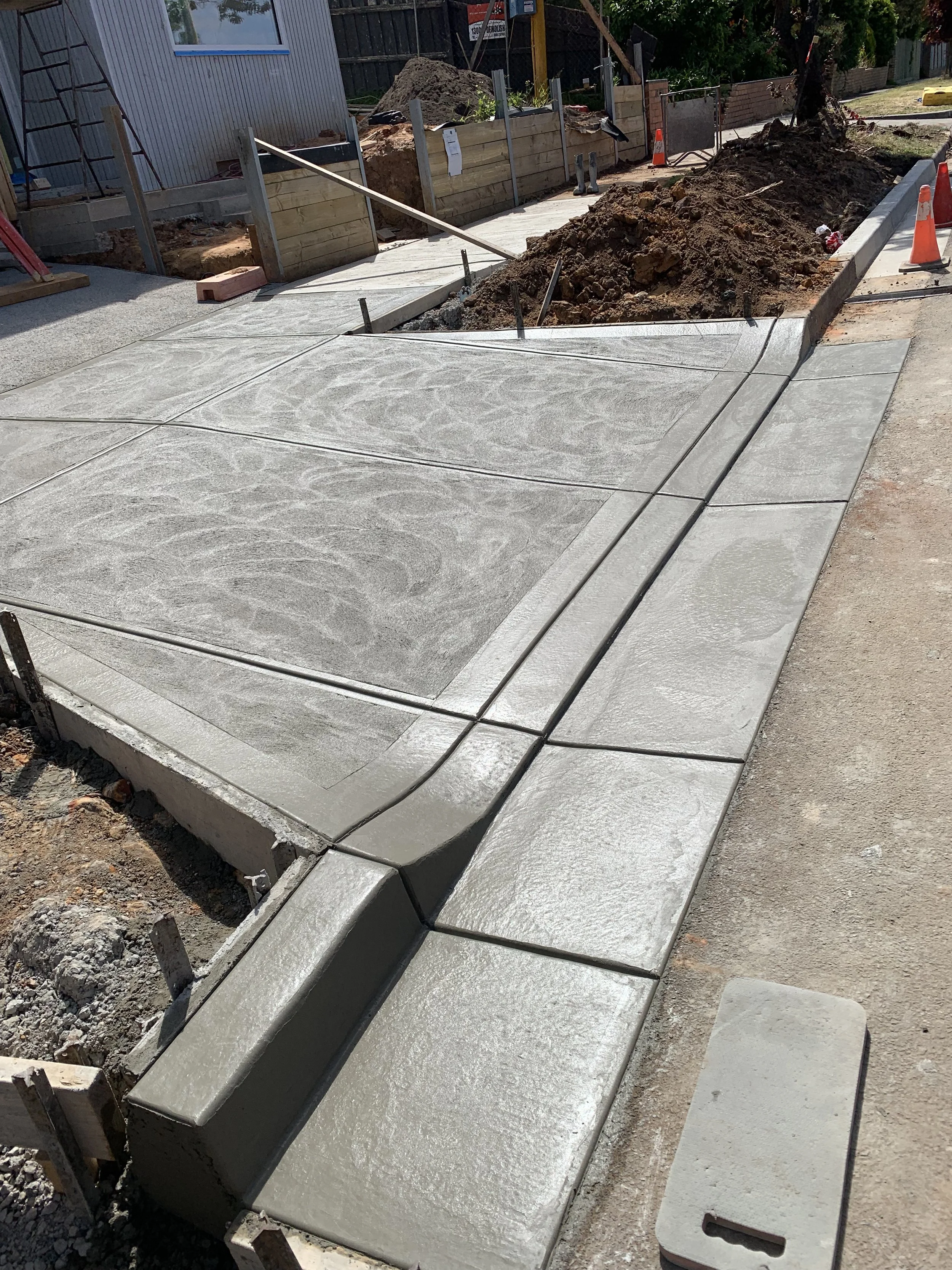 Newly poured concrete sidewalk under construction, with some sections still wet, surrounded by orange safety cones and construction materials. Wooden formwork and dirt piles are visible in the background.
