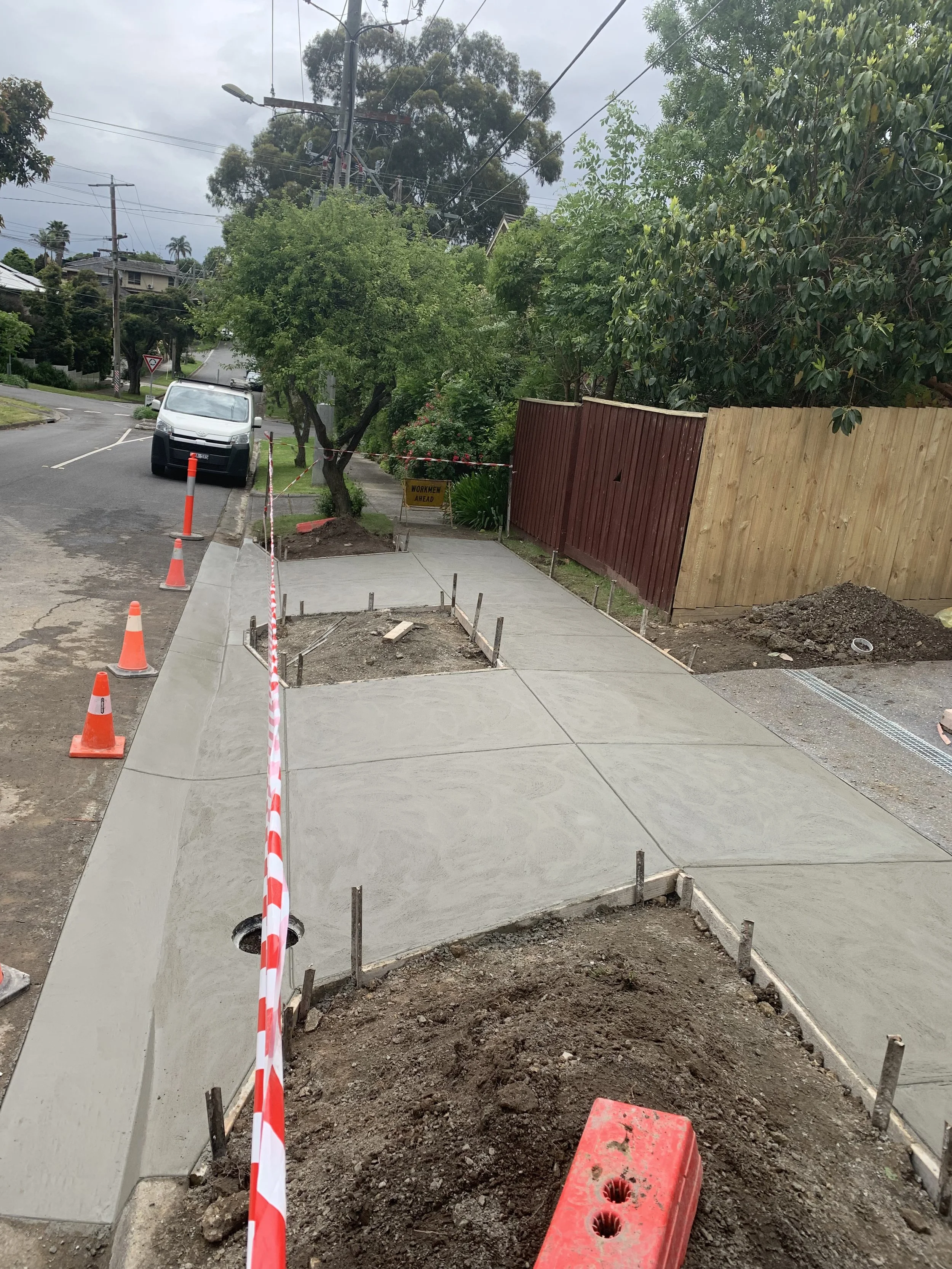 Newly constructed sidewalk with concrete pavement, construction cones, and a warning sign, adjacent to a residential street with trees and fencing.