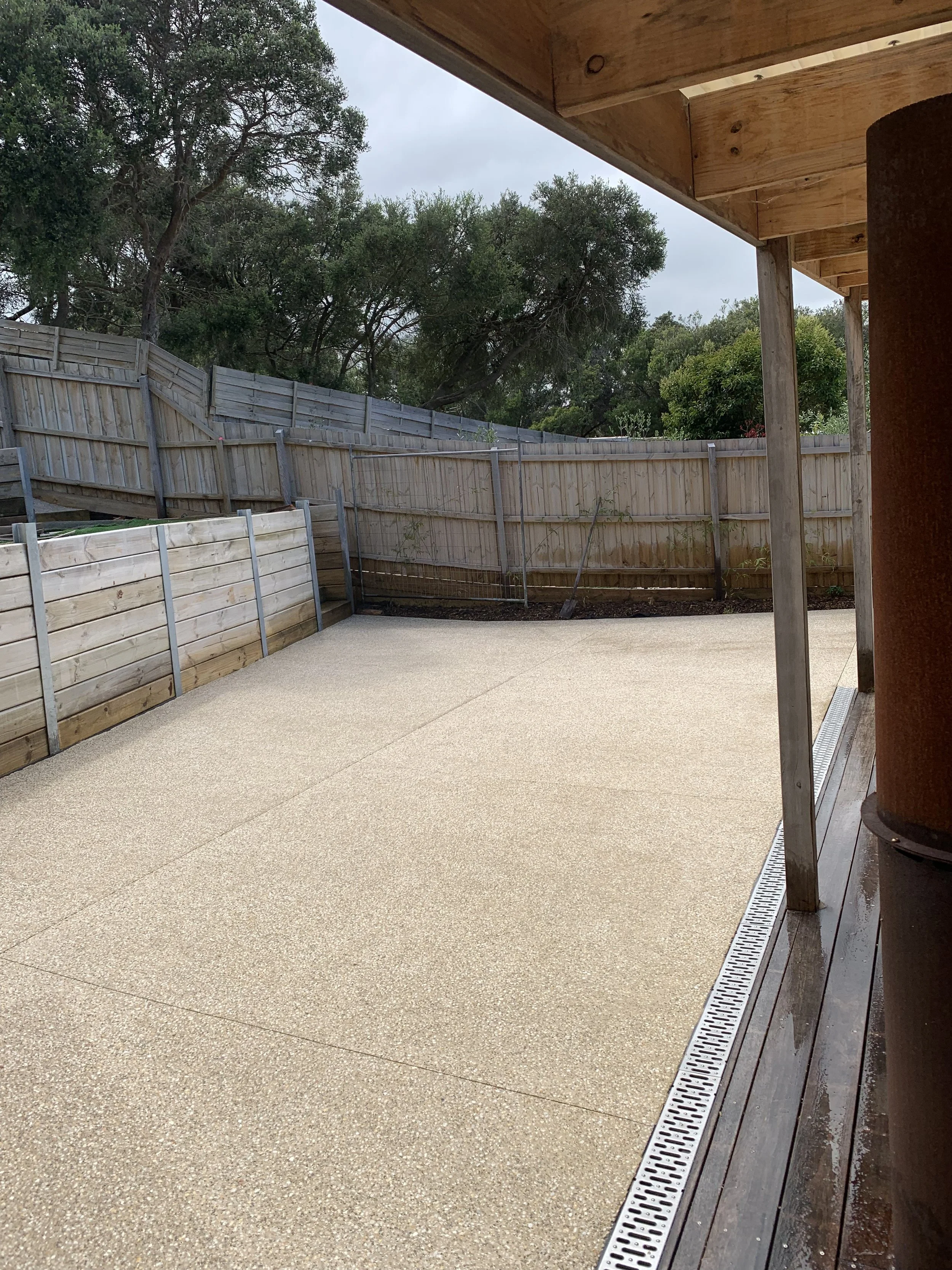 Empty patio with beige textured flooring, wooden fence and trees in the background, partially covered porch with wooden ceiling, and a drainage grate along the edge.