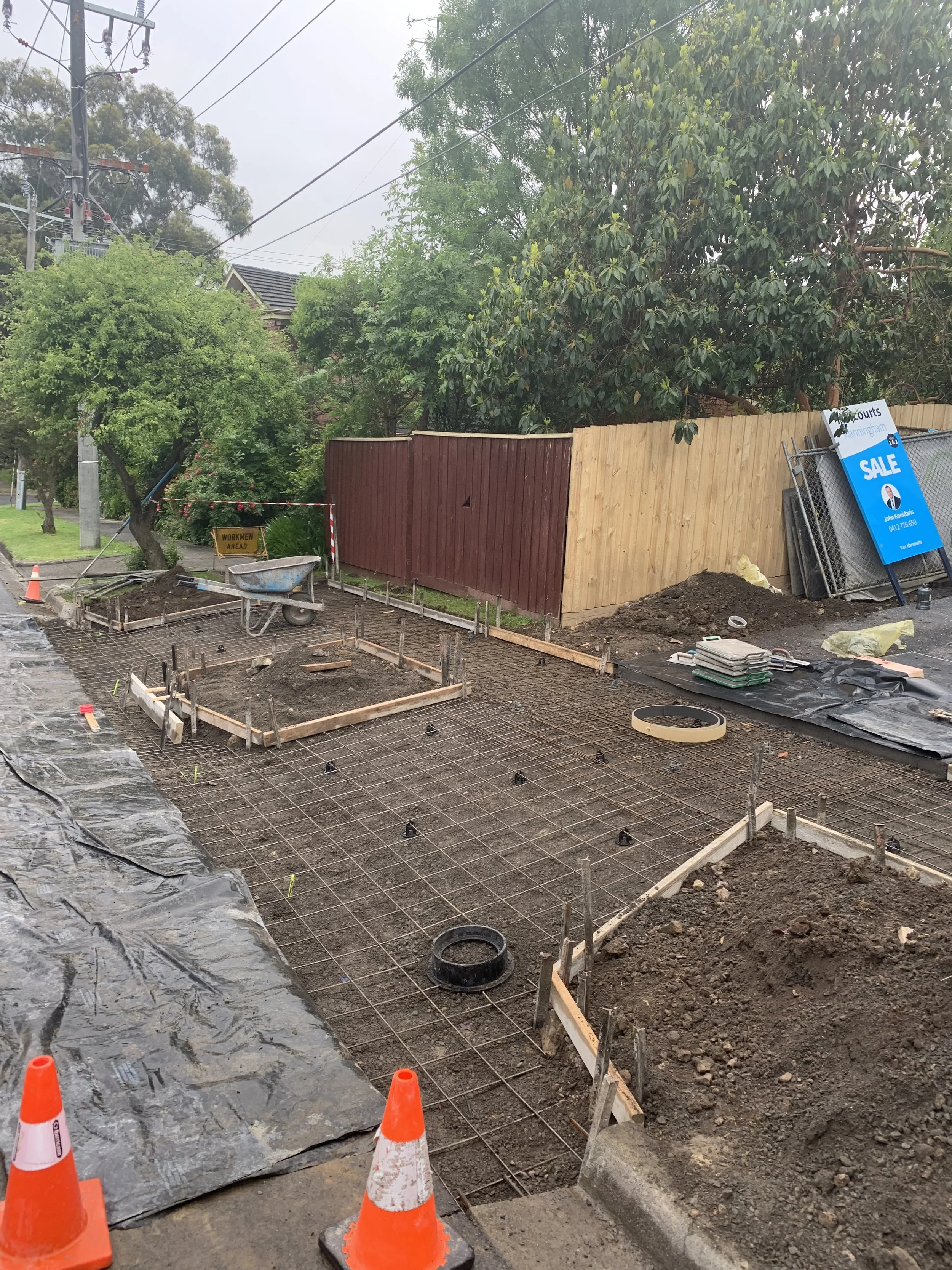 Construction site for a new sidewalk or pavement with orange cones, wooden frames, rebar, and construction tools, with trees, a fence, and a sale sign in the background.