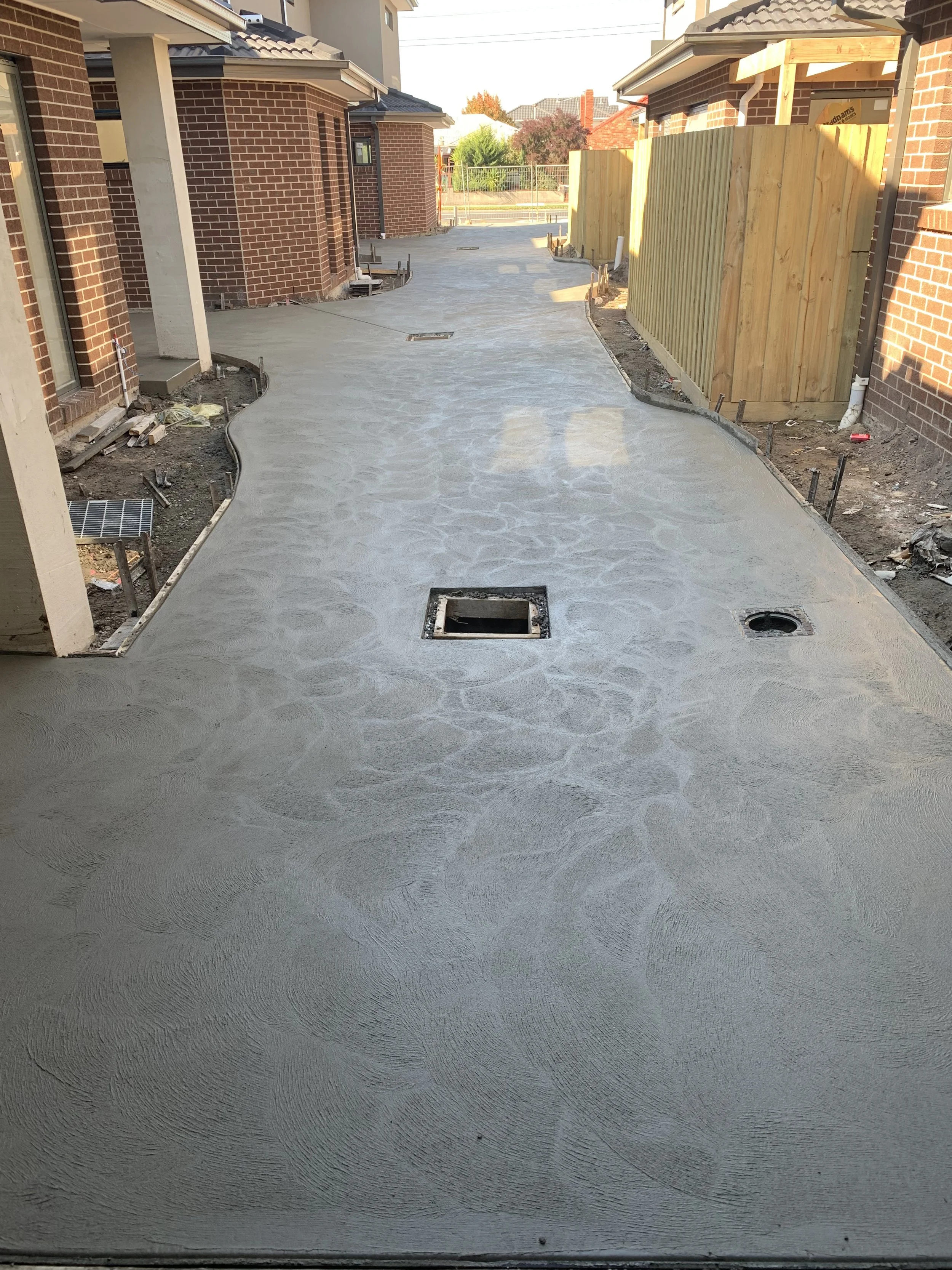 Freshly poured concrete patio with textured finish, surrounded by brick houses and wooden fence, with visible drain openings.