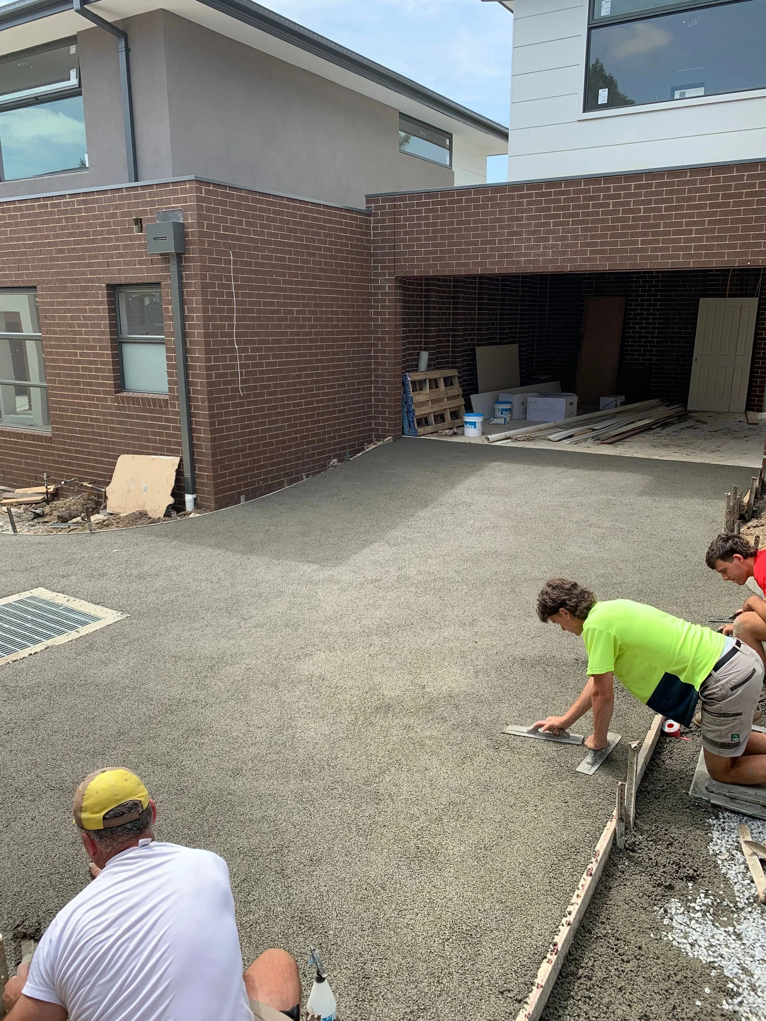 Workers are leveling and smoothing a freshly poured concrete slab outside a modern residential building.