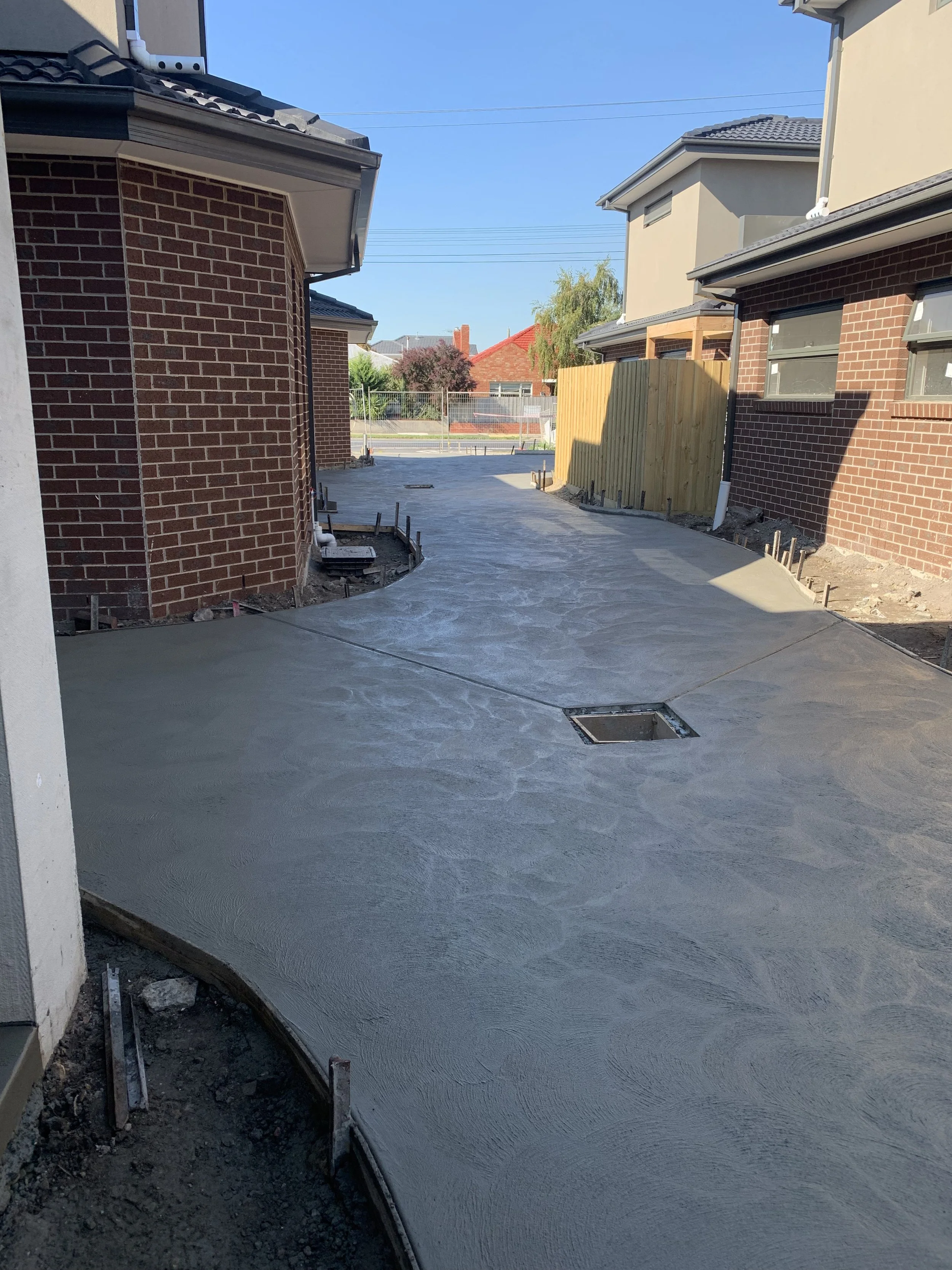 Freshly poured concrete driveway between brick houses under construction, with a wooden fence and neighboring houses visible in the background.