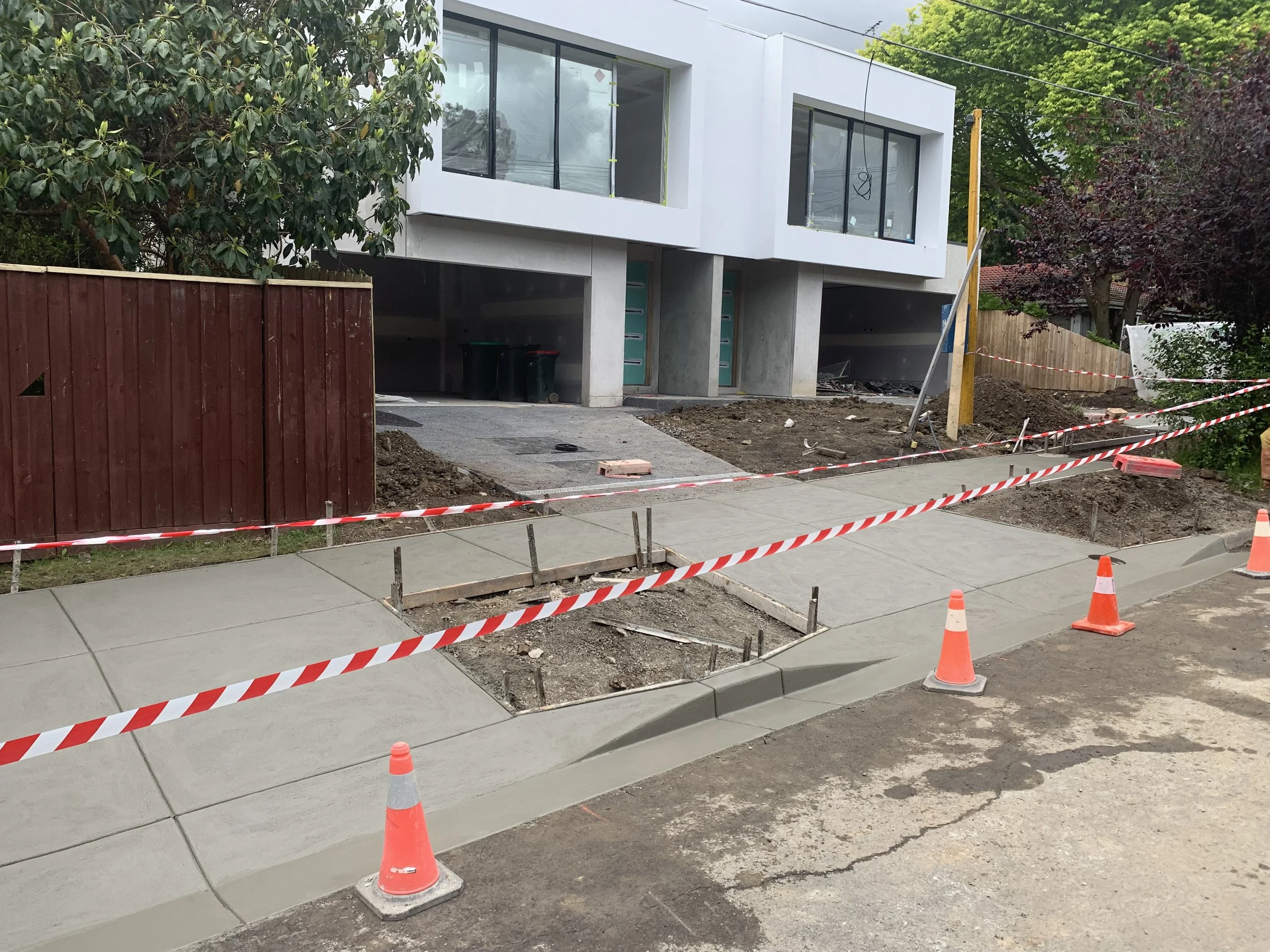Construction of a sidewalk in front of a modern white building with large windows, with orange traffic cones and caution tape surrounding the work area.