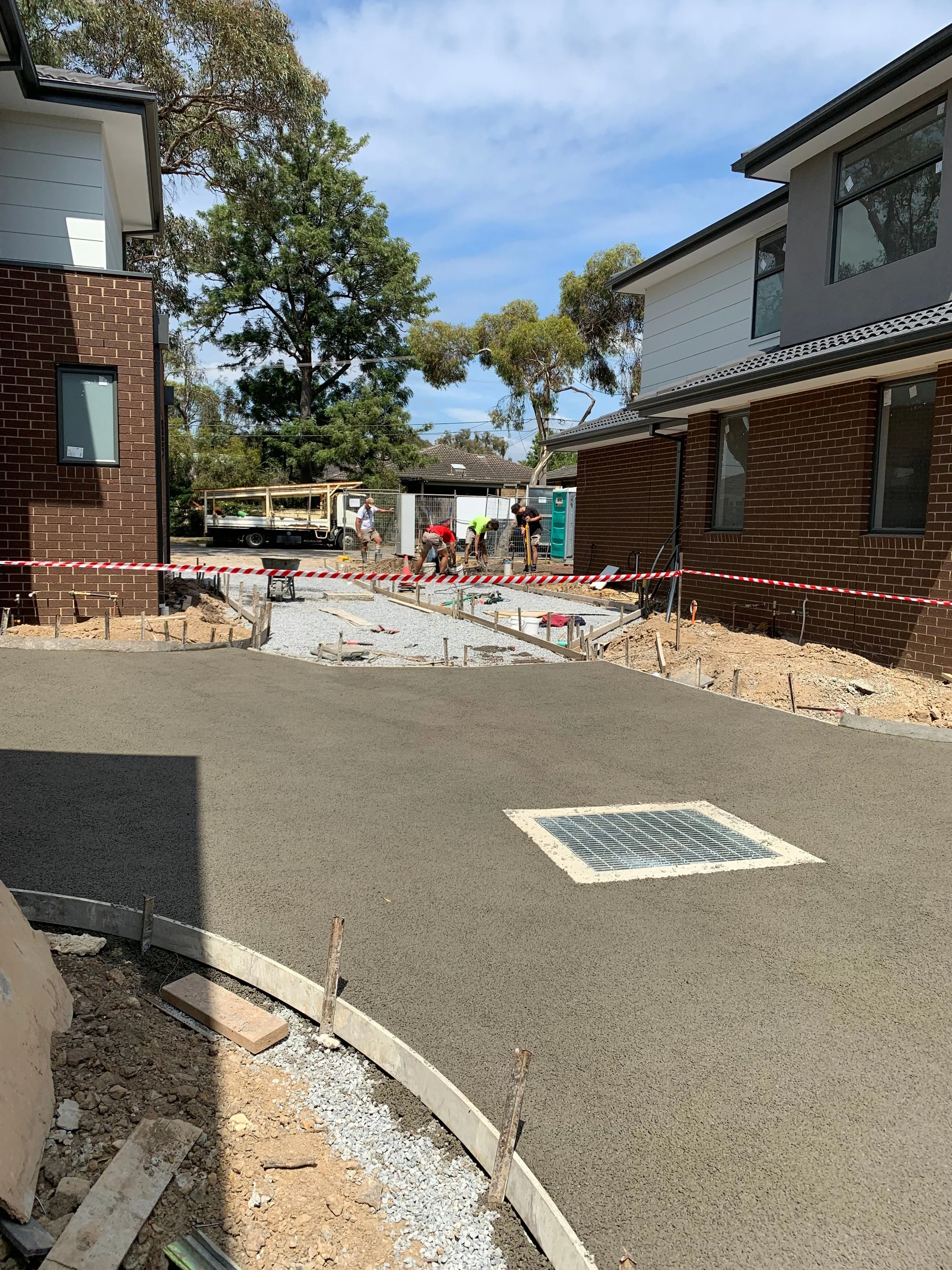 Construction workers laying fresh asphalt on a driveway between two modern residential buildings, with trees and a partly cloudy sky in the background.