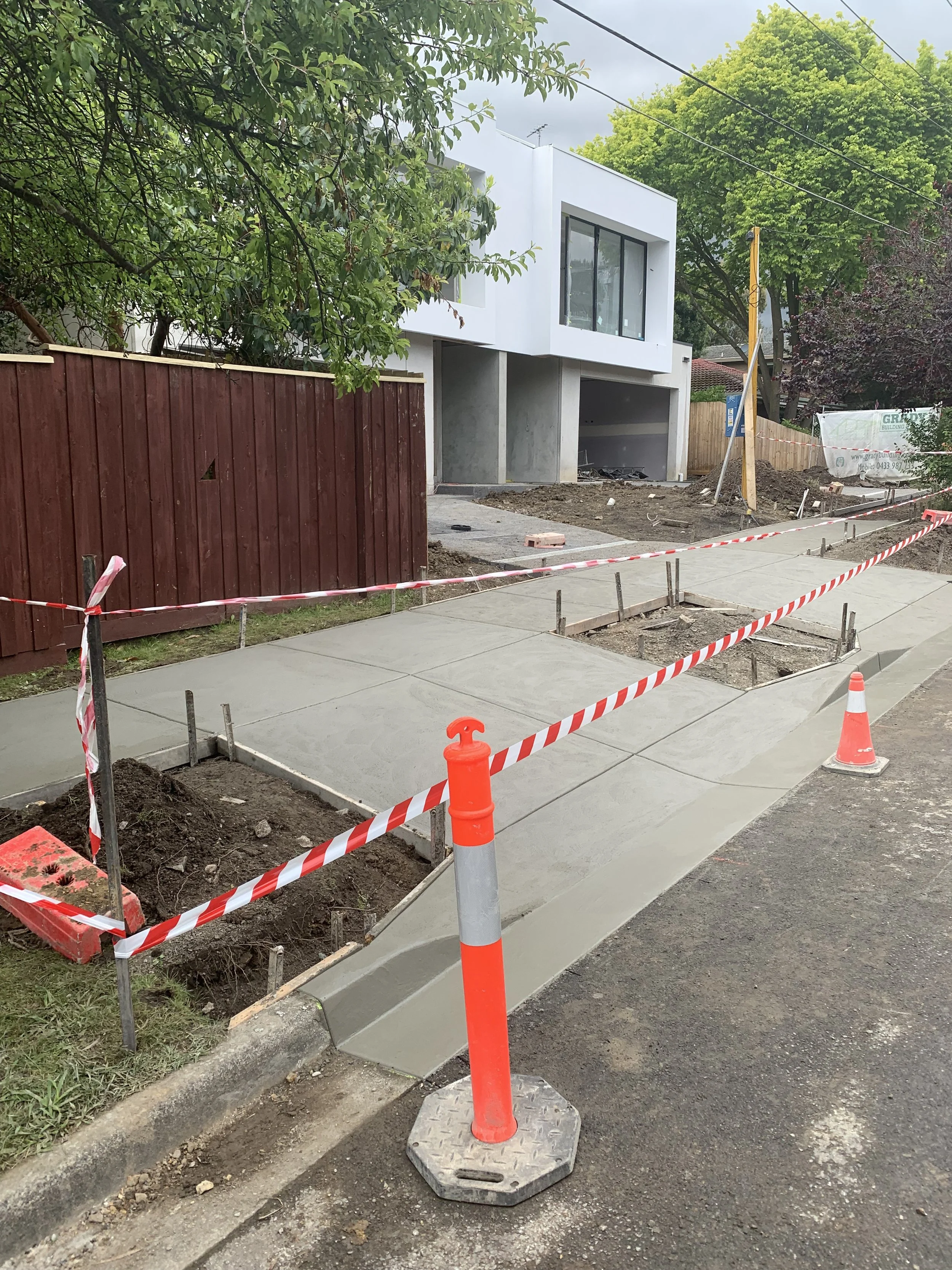 Construction site with new concrete sidewalk and curb, orange safety cones and barriers, in front of a modern white building under construction.