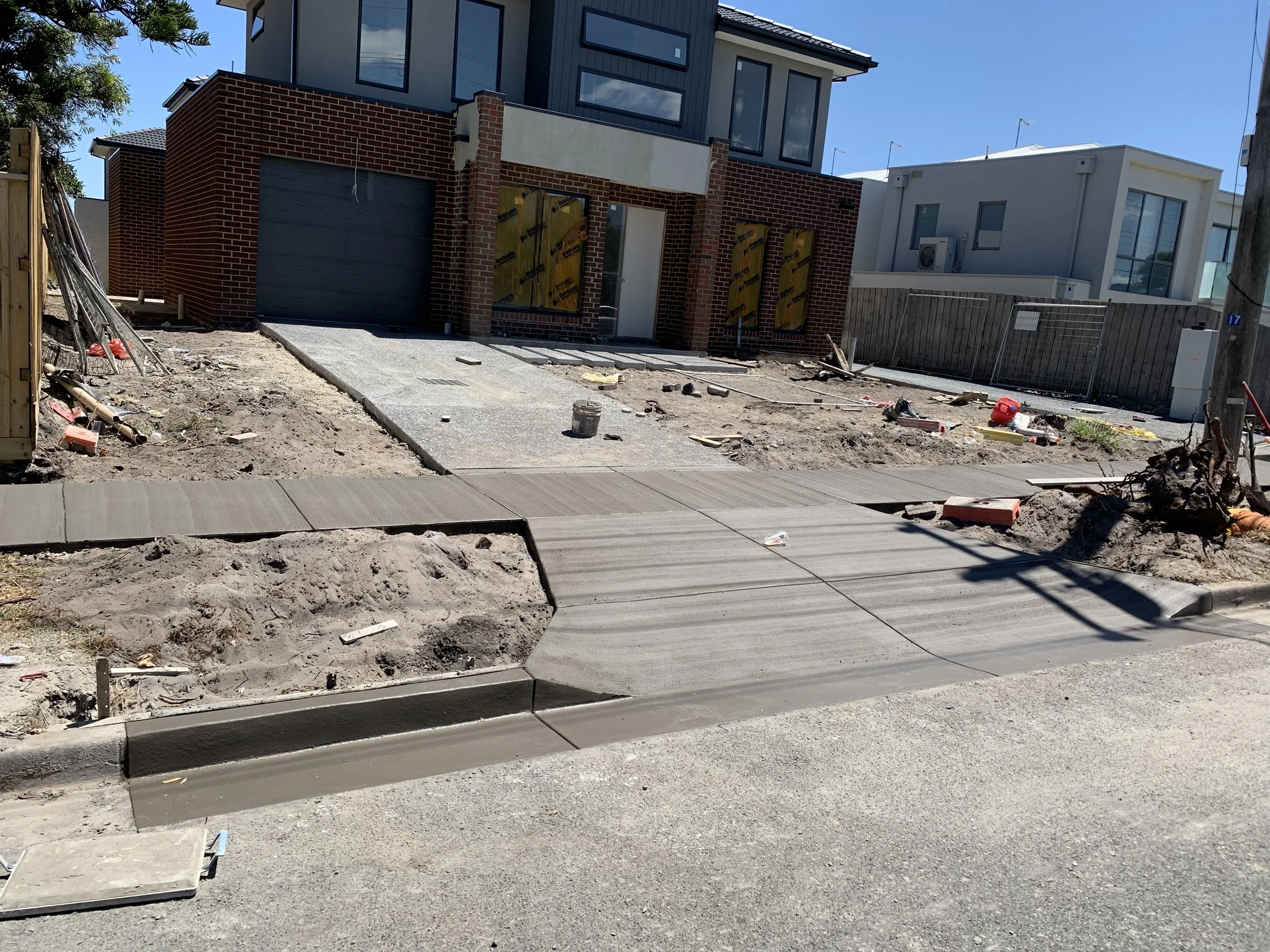 Under construction house with new concrete sidewalk in front, scattered construction tools and debris, and modern building in the background.
