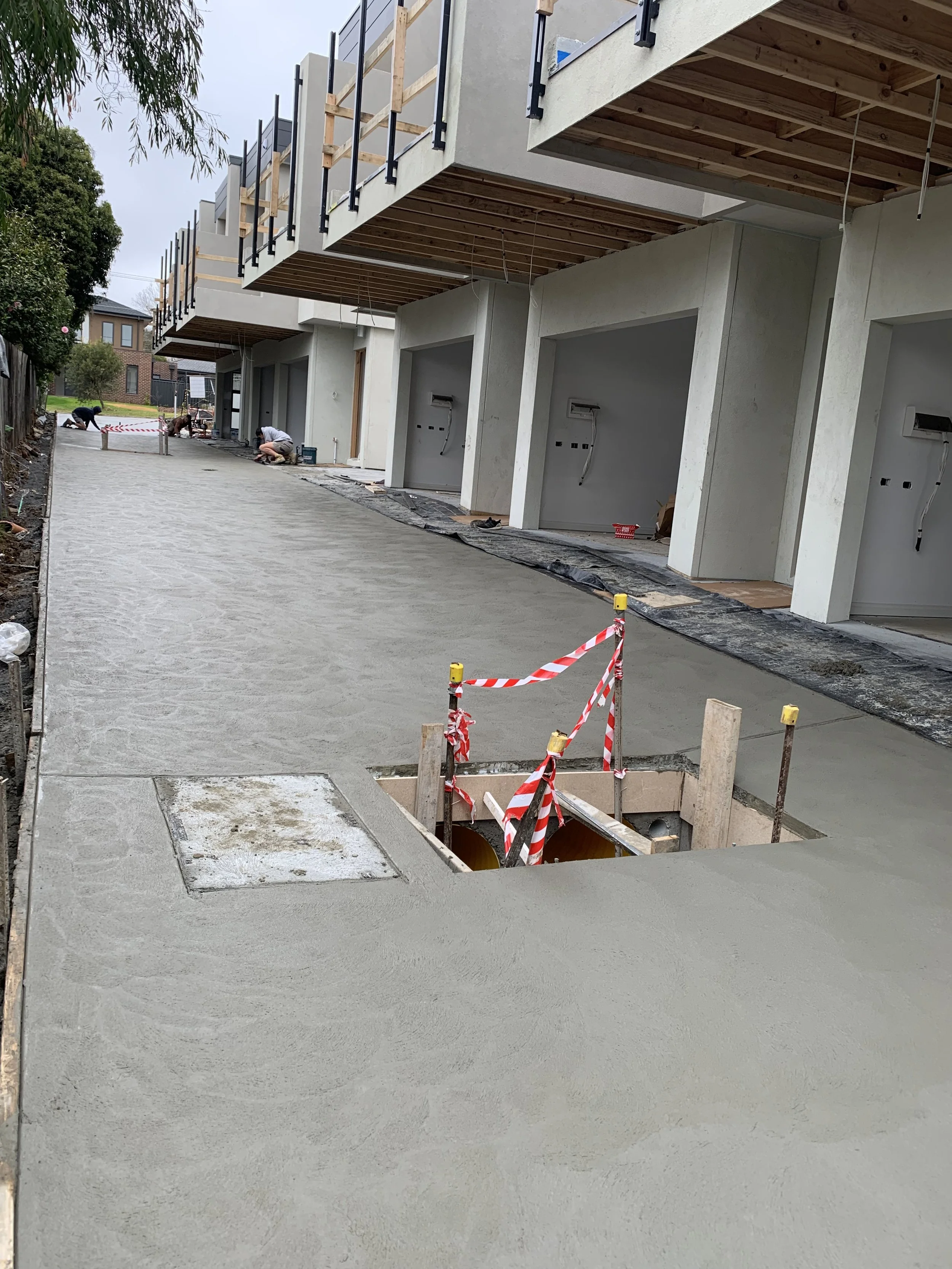 Newly constructed apartment complex with a concrete walkway, some garage openings, and ongoing work. Construction workers are seen in the background, and there is a safety barrier around an open utility hole in the foreground.