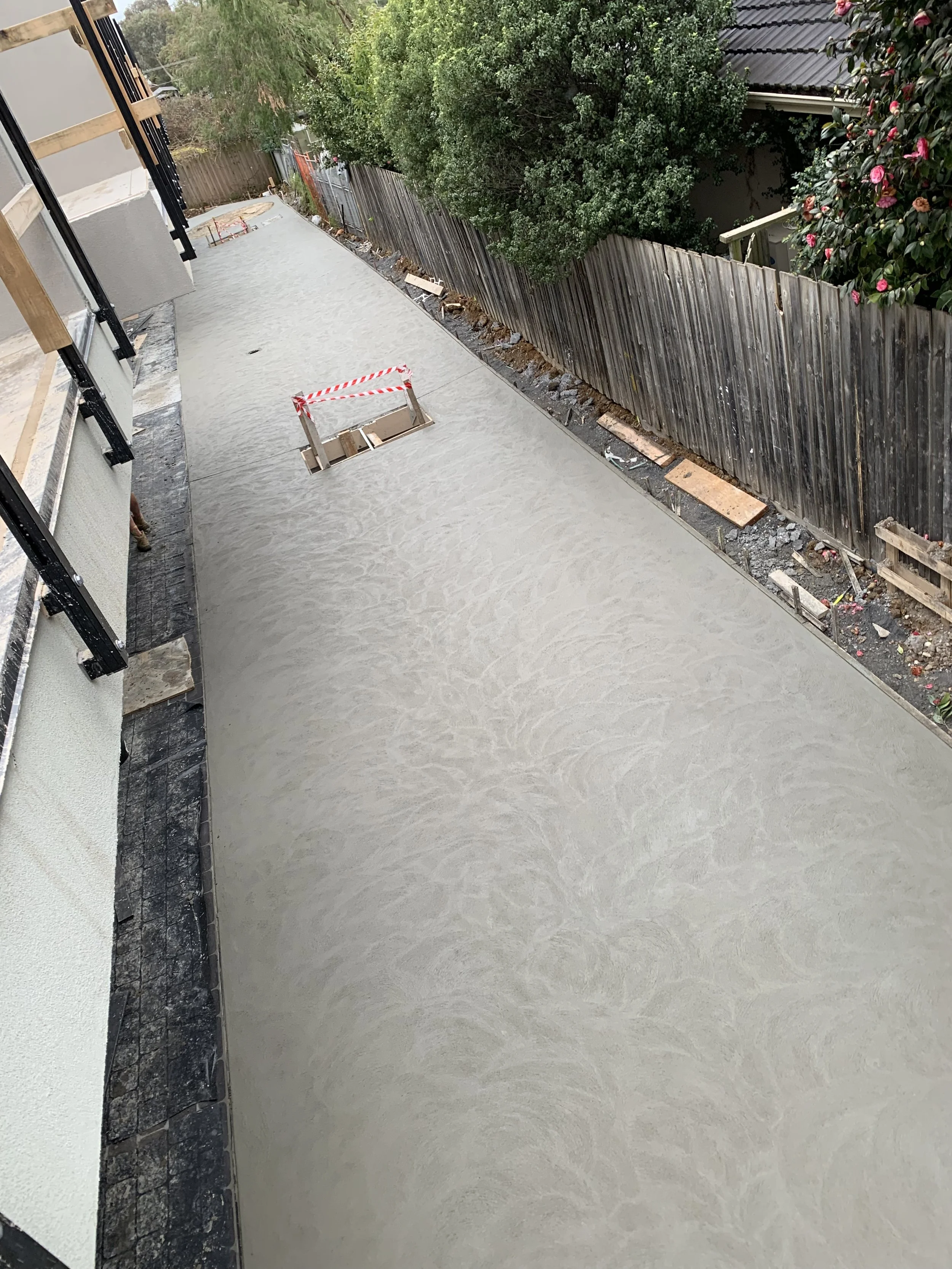 A freshly poured concrete pathway under construction next to a building with balcony and a wooden fence, with trees and pink flowering bushes in the background.