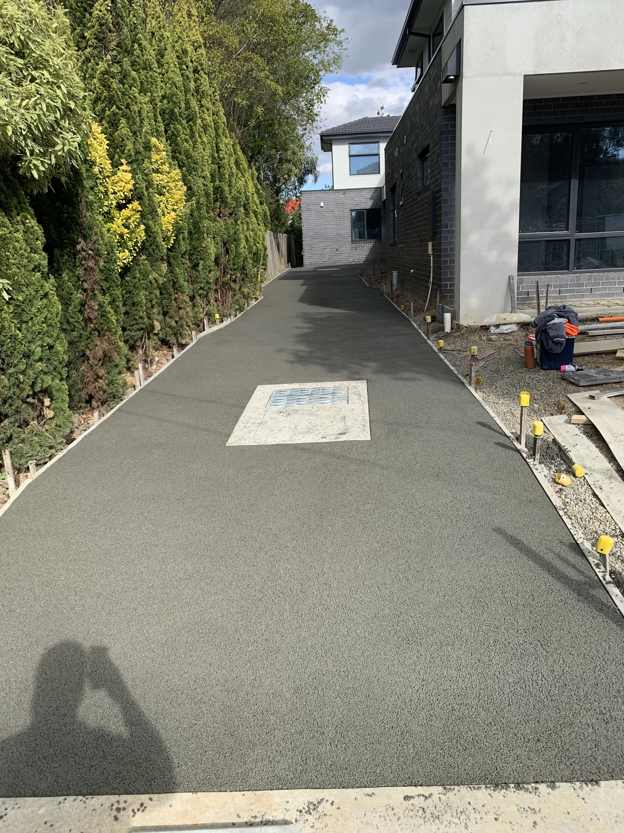 Newly paved concrete pathway leading up to a modern residential building, with construction materials and tools on the side, and green trees lining the left side.