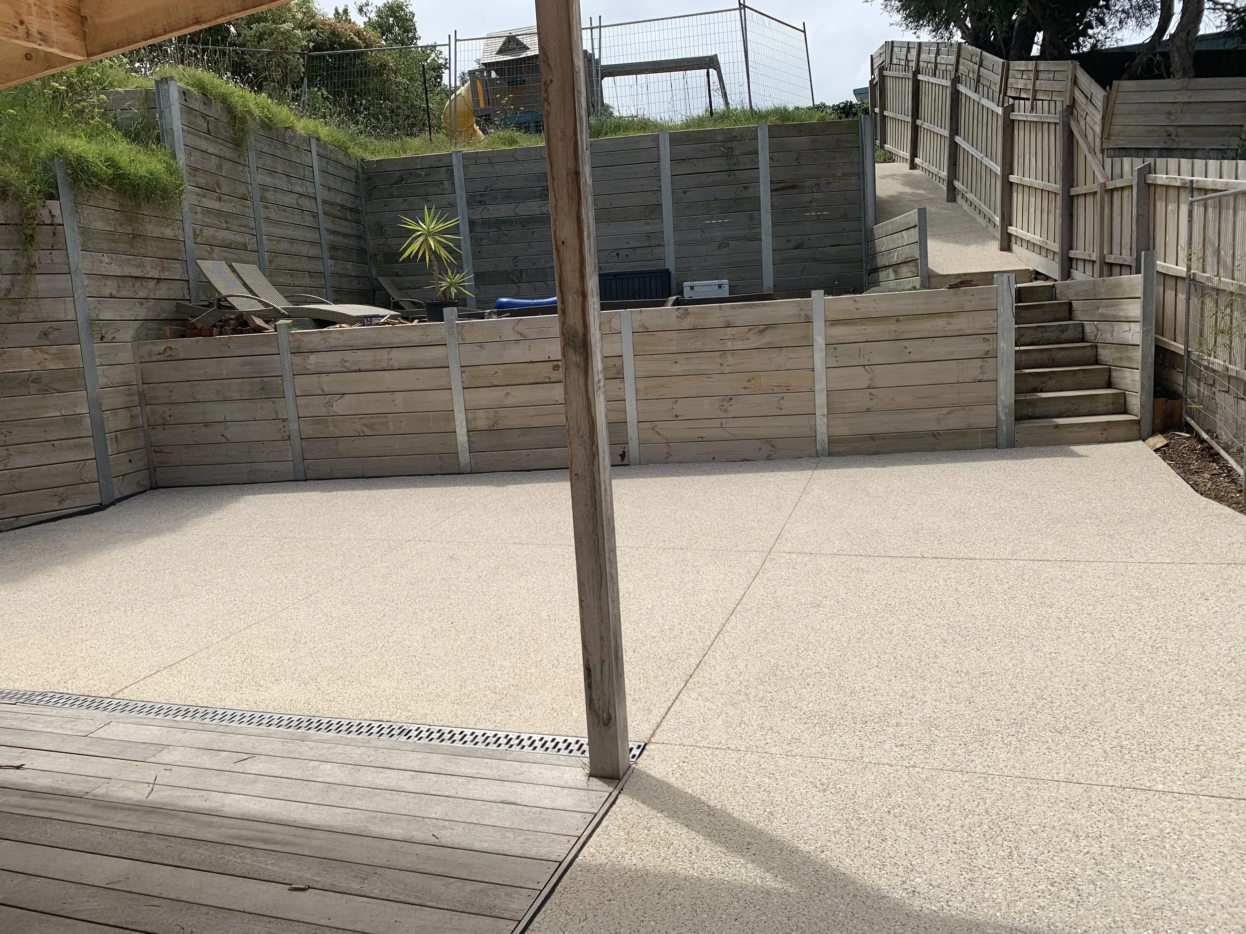 A backyard with a concrete patio, wooden stairs leading to a higher level, and wooden fences surrounding the yard. There are some lounge chairs and a potted plant on the upper level.