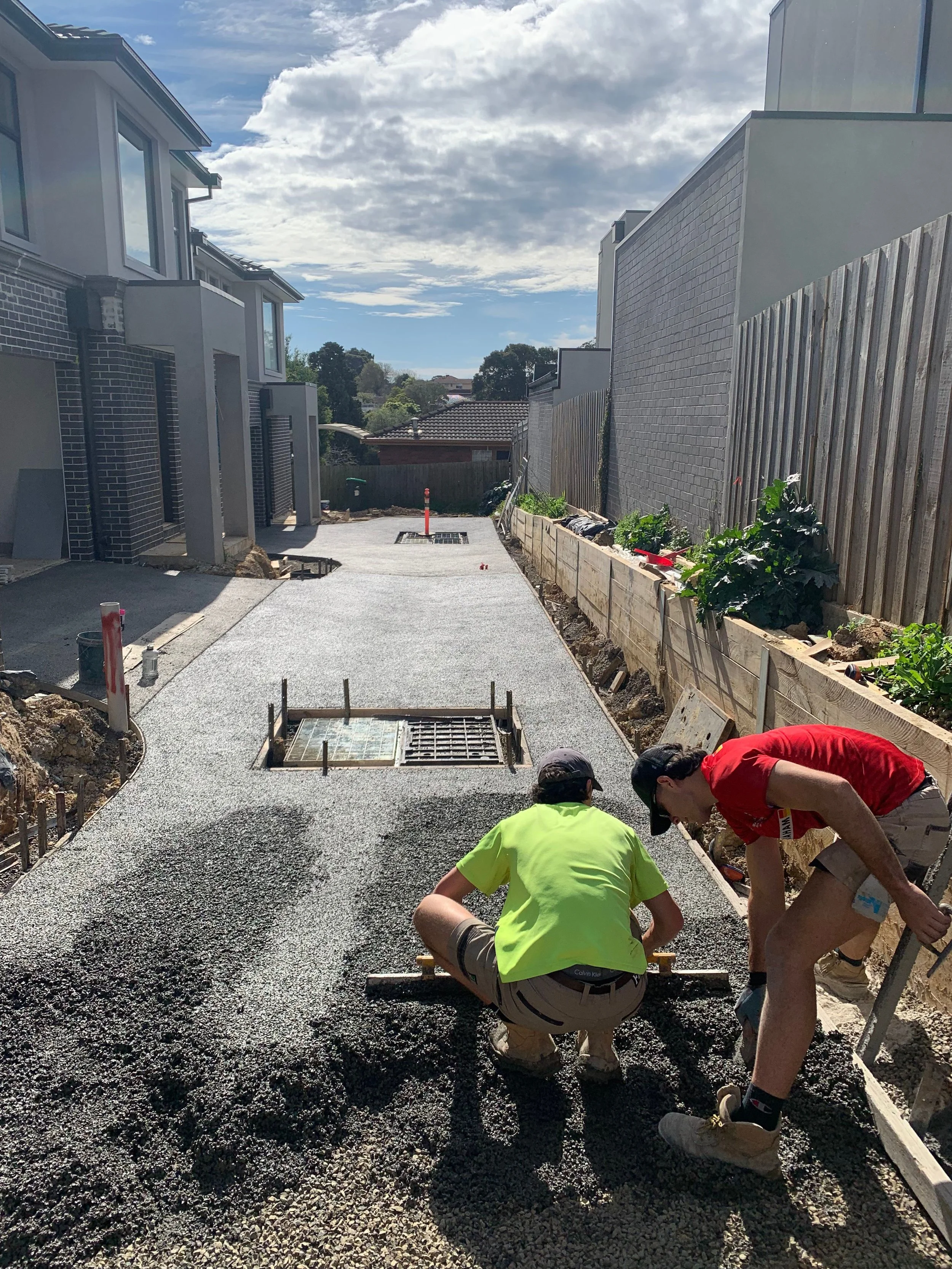 Workers pouring and leveling freshly laid asphalt on a driveway in a residential area under a partly cloudy sky.