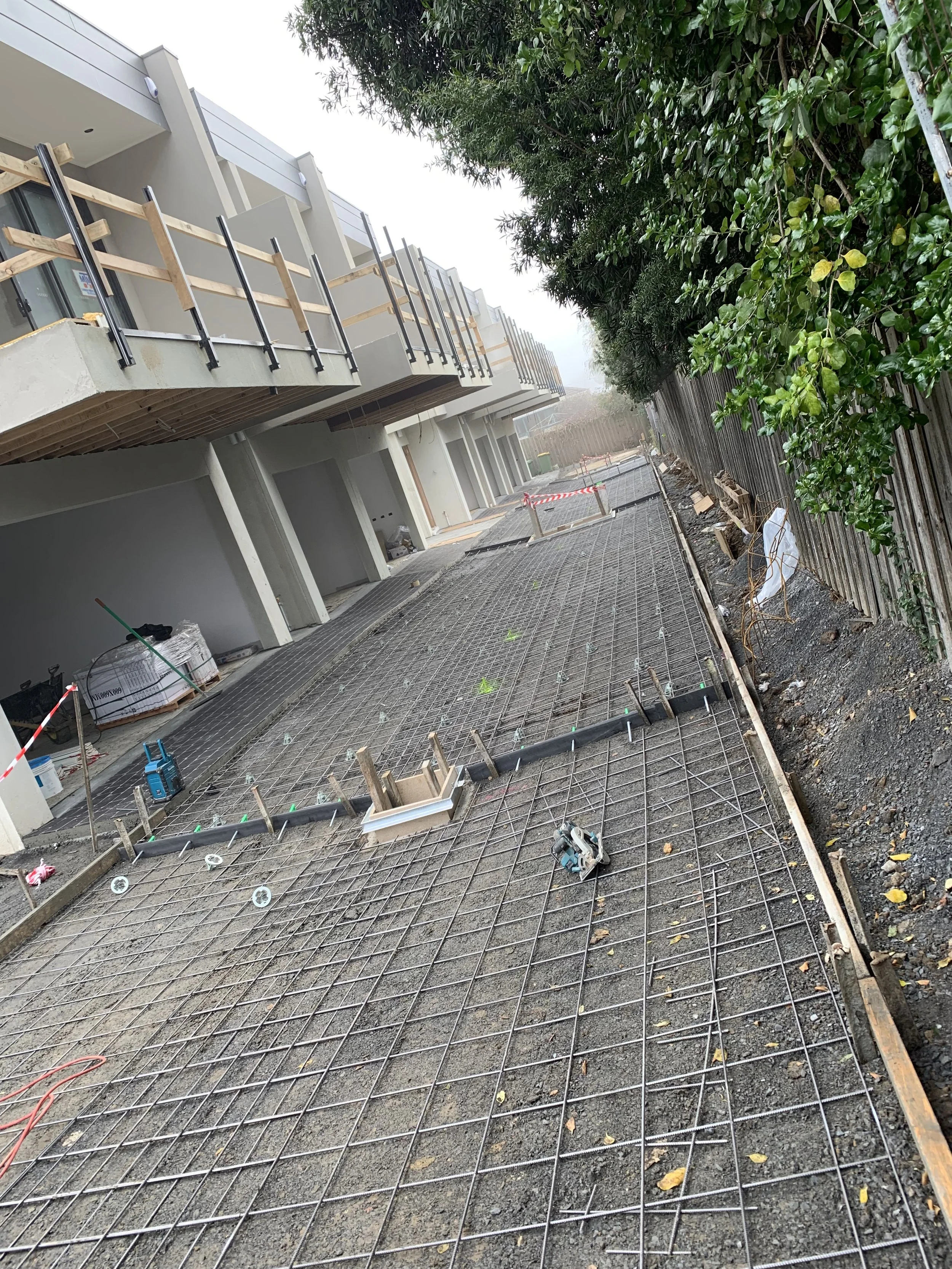 Under construction sidewalk with reinforcement steel mesh, nearby building with balconies, trees, and a fence.