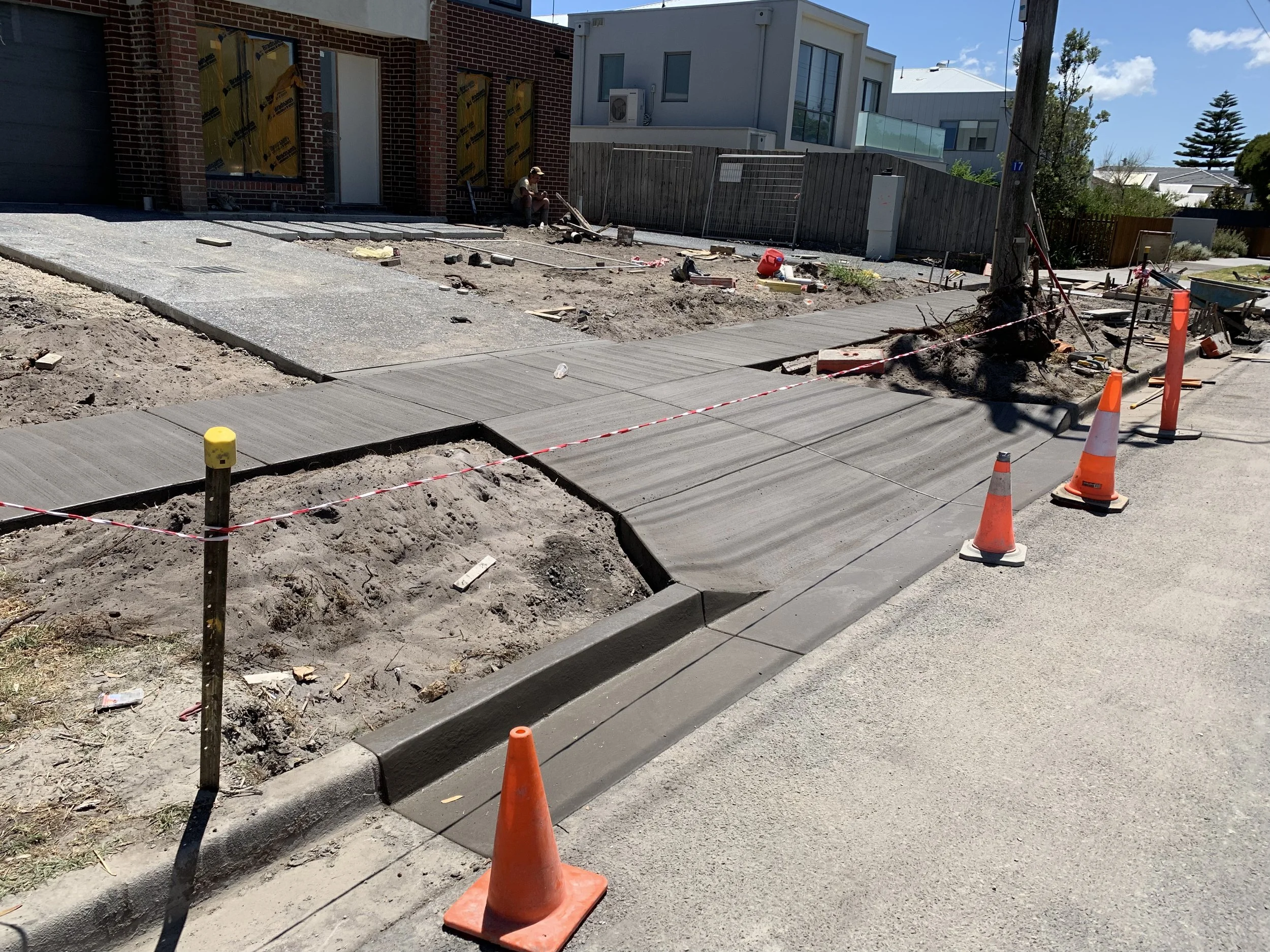Construction site with newly poured concrete sidewalk, orange cones, and a tree surrounded by construction materials. House under renovation in the background.