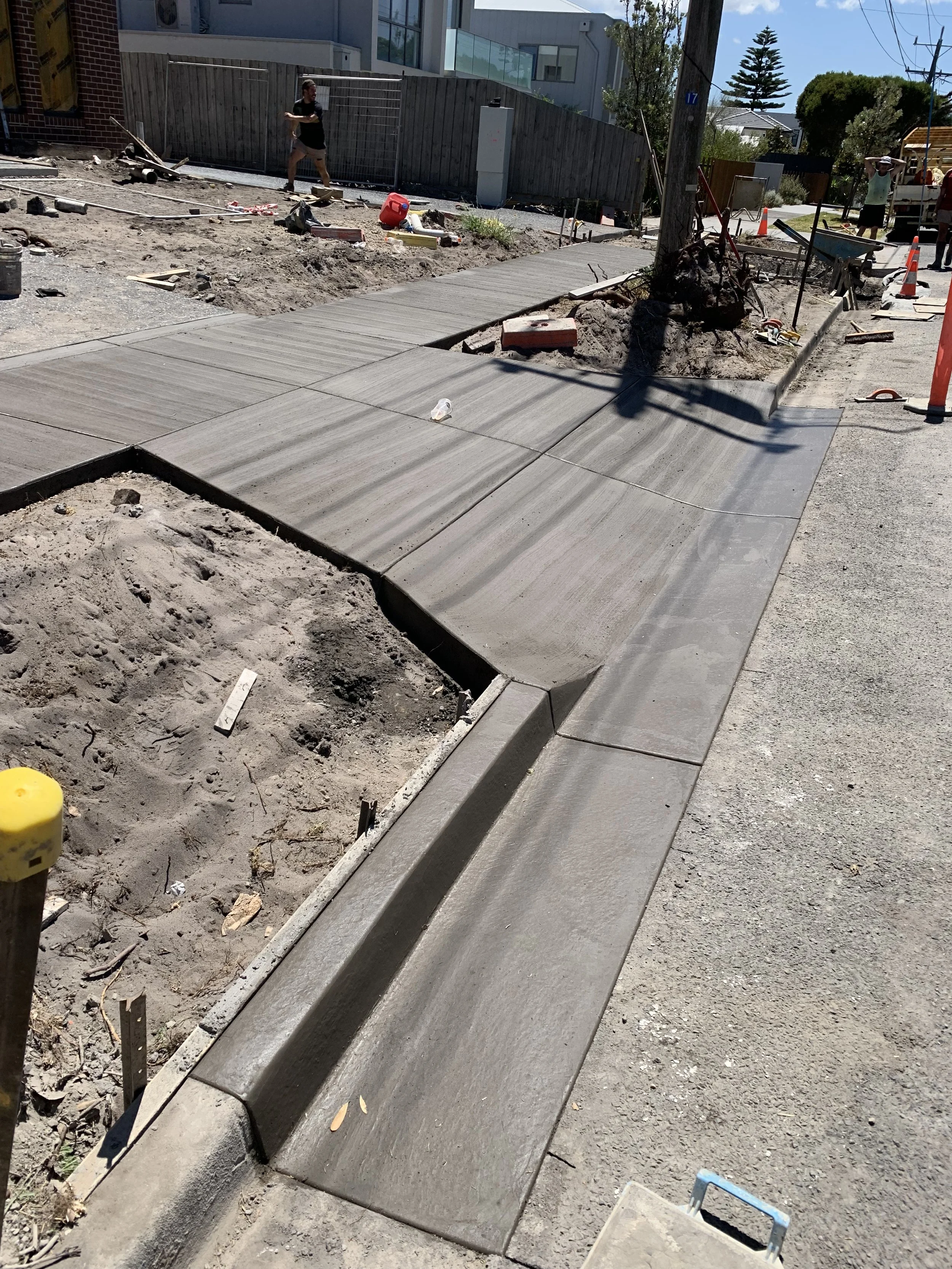 Construction site with a newly laid concrete sidewalk, a dug-up area on the left, construction tools and materials scattered around, and some workers in the background.
